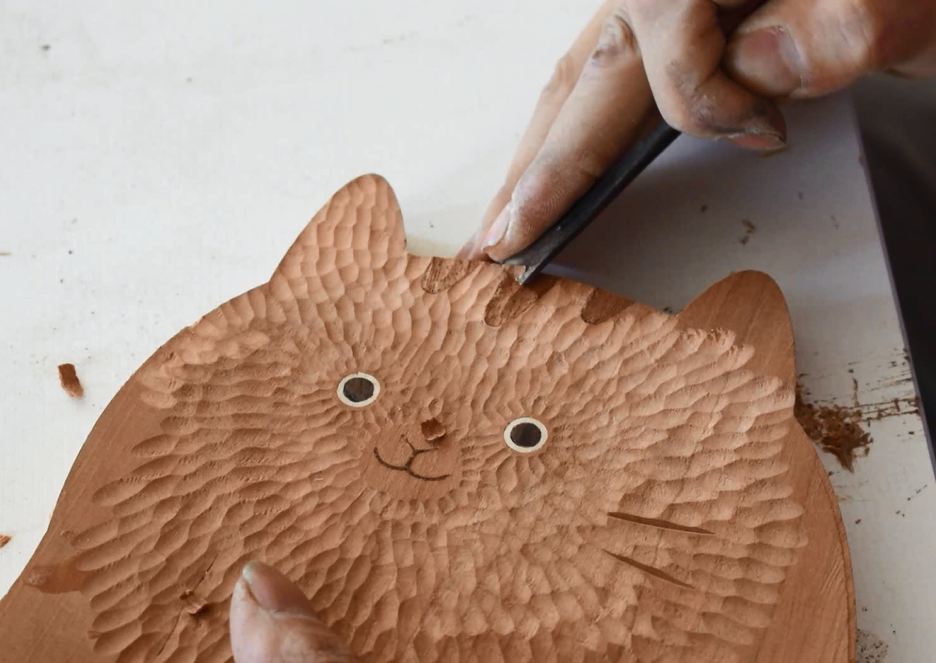 Wooden cat figurine being carved with a tool on a white surface
