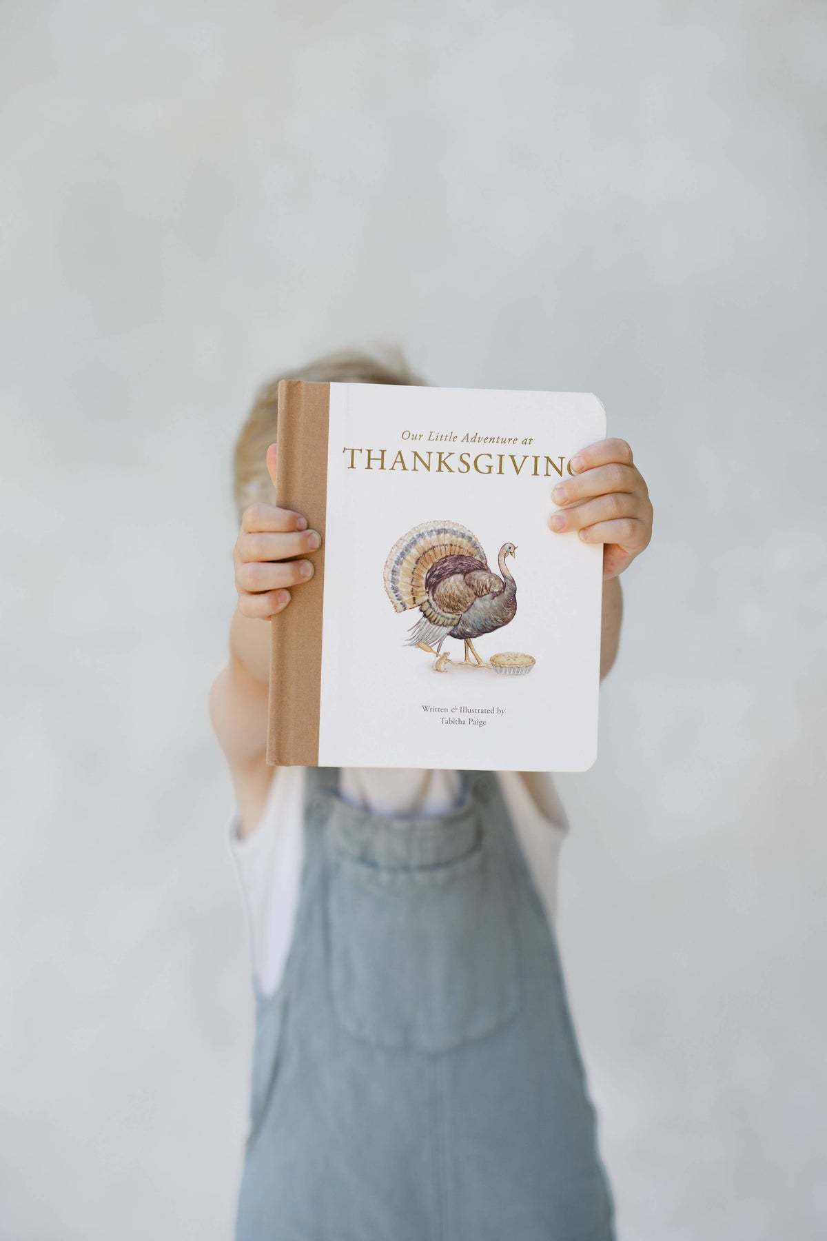 A child holds up a hardcover children's book titled 'The Little Adventure of Thanksgiving' against a light gray background. The child is wearing blue denim overalls and a white shirt, with only the hands and lower body visible.