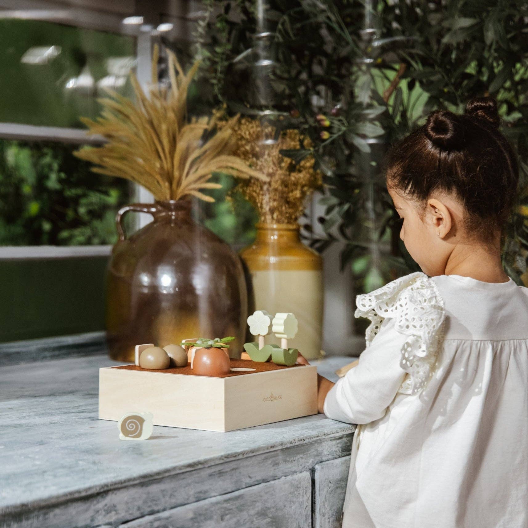 Child standing near a wooden dresser with decorative items and plants.