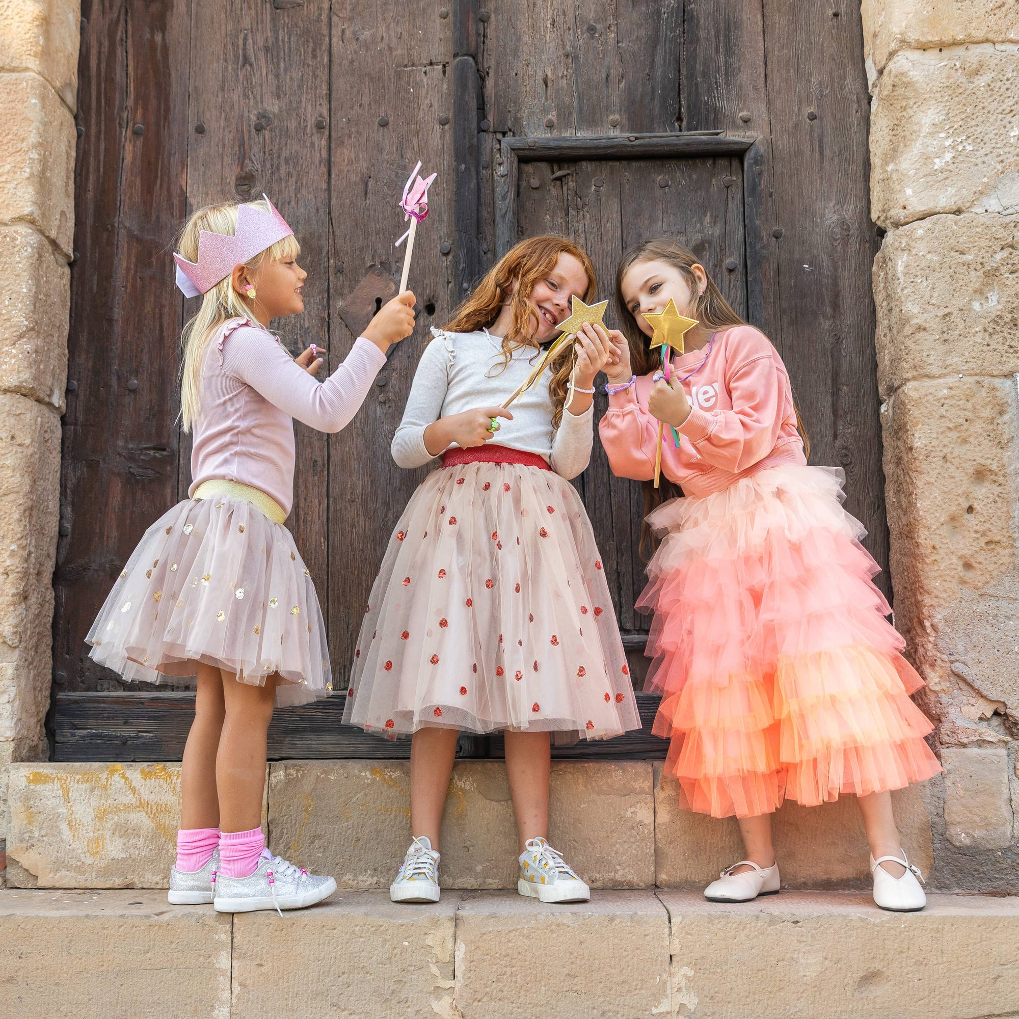 Three young girls in colorful dresses standing in front of a large wooden door.