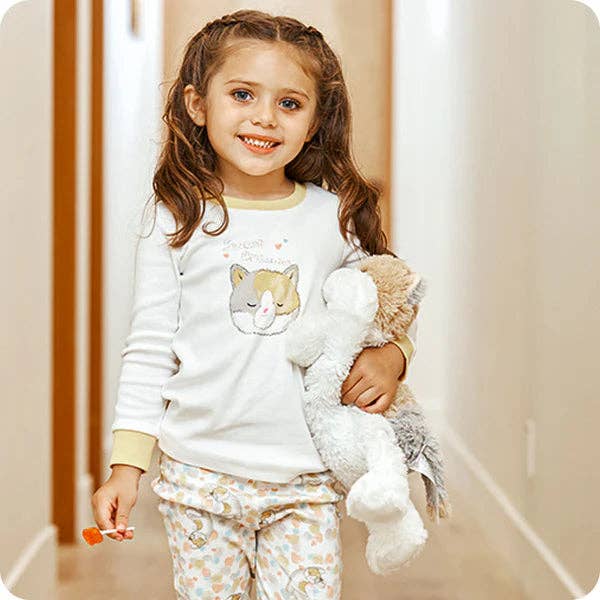 Young girl in pajamas holding a teddy bear and a small toy, standing in a softly lit room.