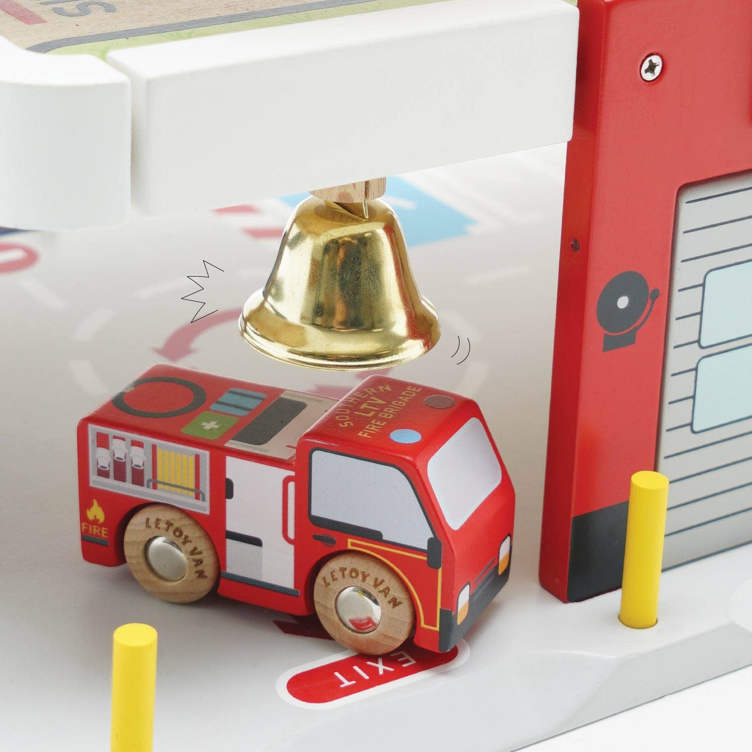 Toy fire truck with a bell on top, next to a toy fire station, on a white surface.