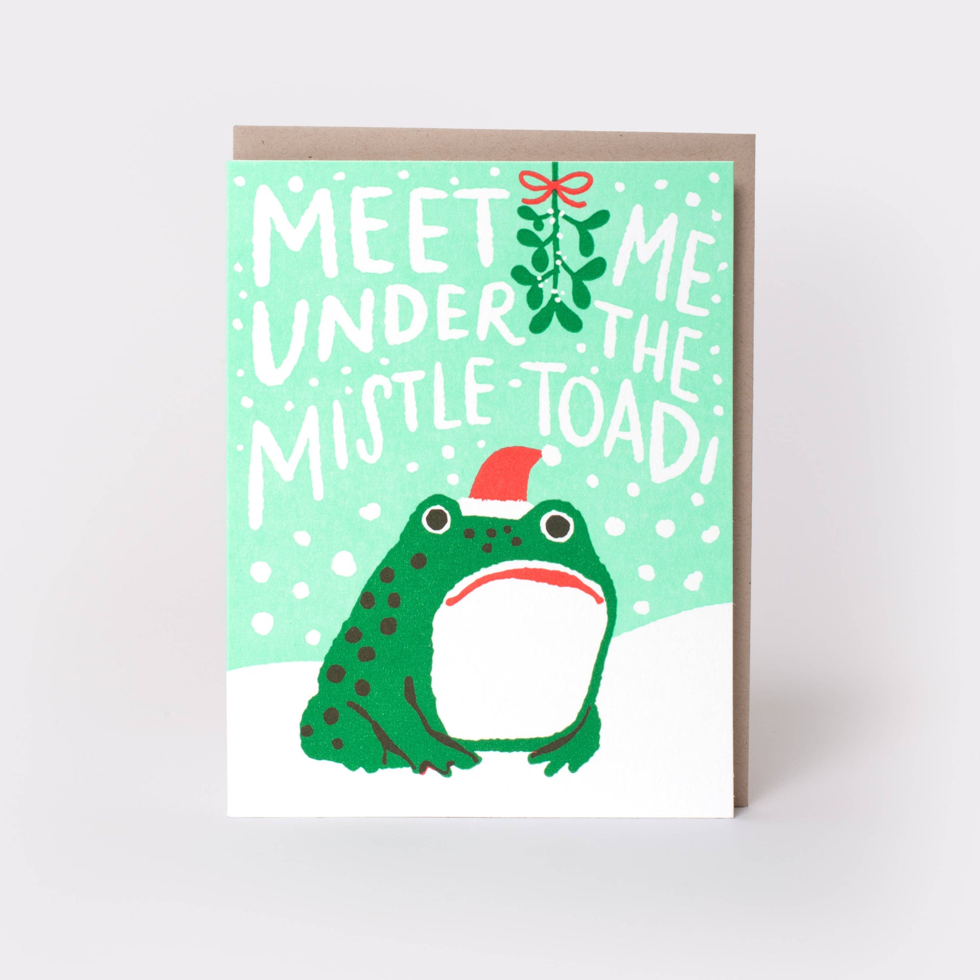 A Christmas greeting card featuring a green toad wearing a red Santa hat, with the text 'MEET ME UNDER THE MISTLE TOAD!' and a mistletoe sprig on a mint green background. The card is shown standing upright with a brown envelope behind it on a light gray background.