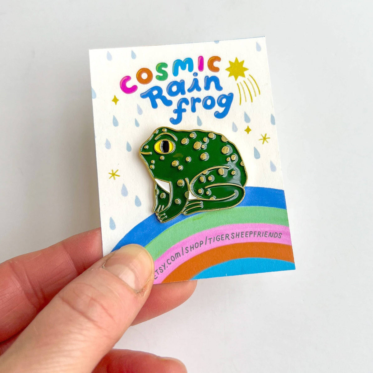 Pin with a green frog design on a card labeled 'Cosmic Rain Frog' held by a hand.