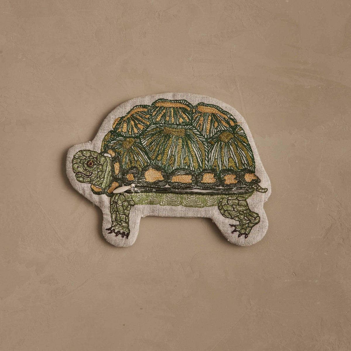 An embroidered patch shaped like a tortoise, featuring green and yellow-green stitching with tan accents on a light beige fabric background, displayed on a textured beige surface.