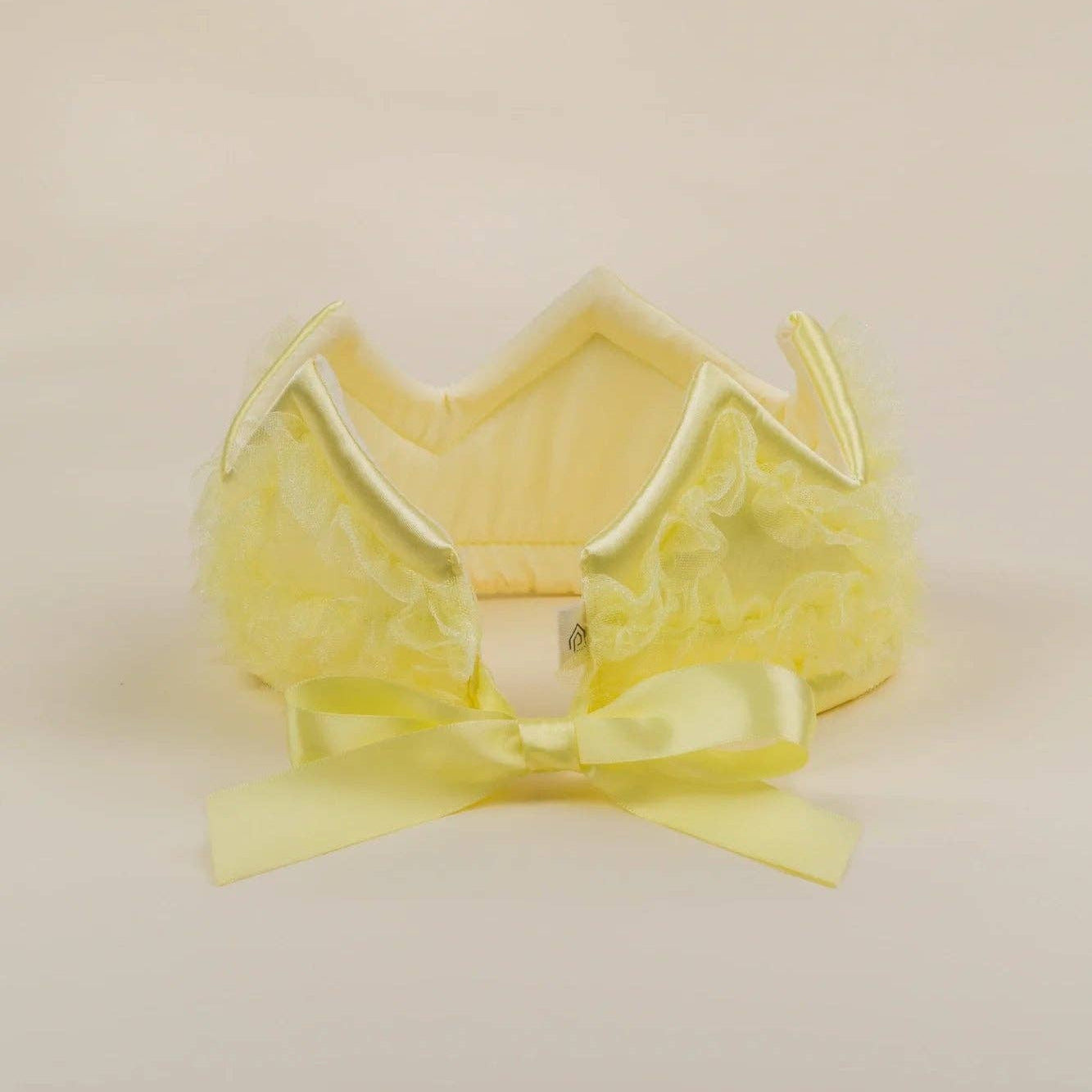 A pale yellow lace and tulle crown with a matching satin bow, displayed on a neutral cream background.