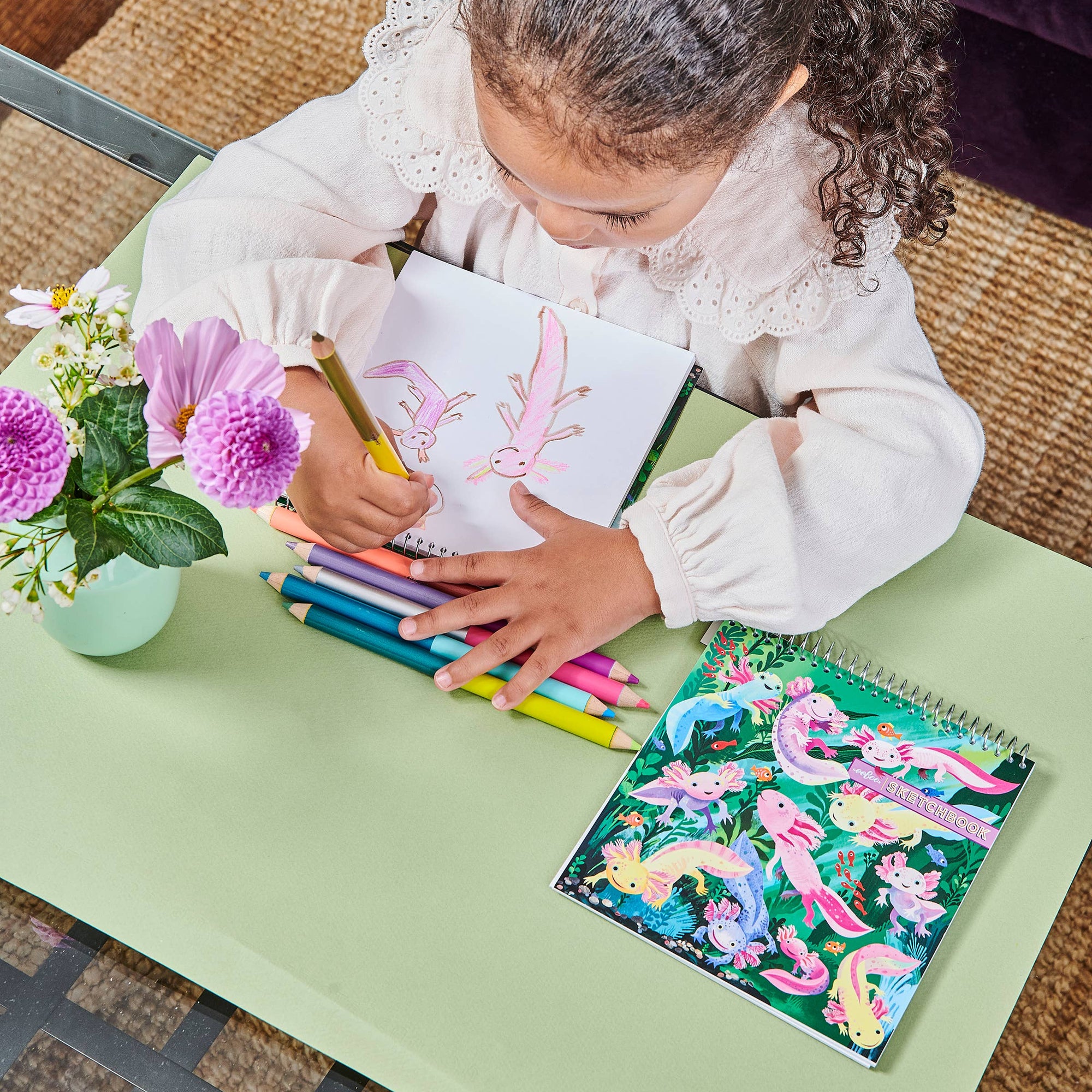Child coloring with crayons on a table with coloring books and flowers.