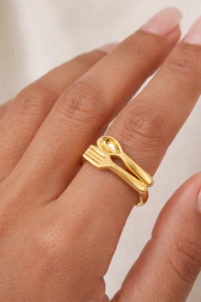 Gold ring with fork and spoon design on a hand against a neutral background