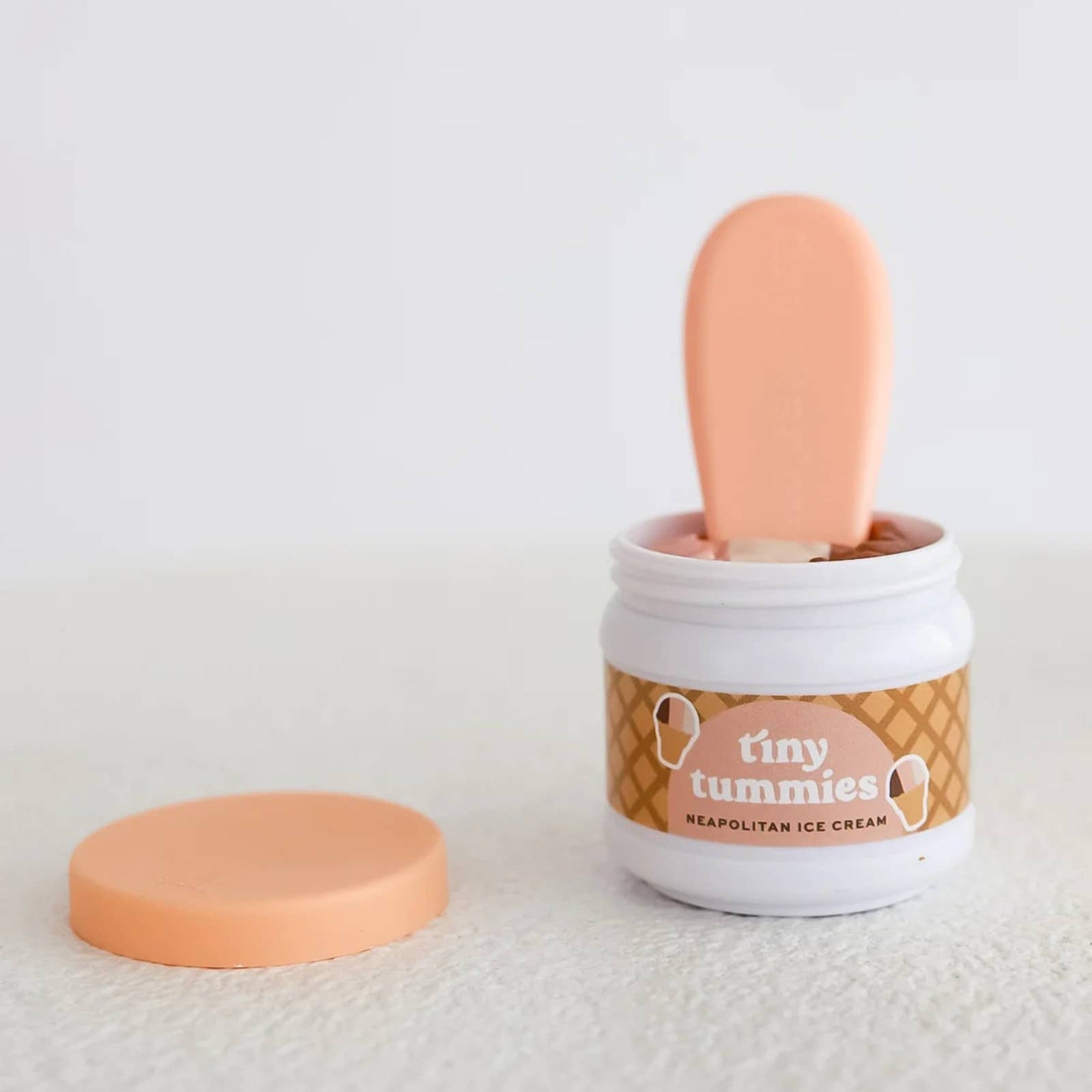 A small white jar of 'Tiny Tummies' whipped ice cream with a peach-colored scoop resting inside, shown with its matching peach lid placed beside it on a soft white textured surface against a clean white background.