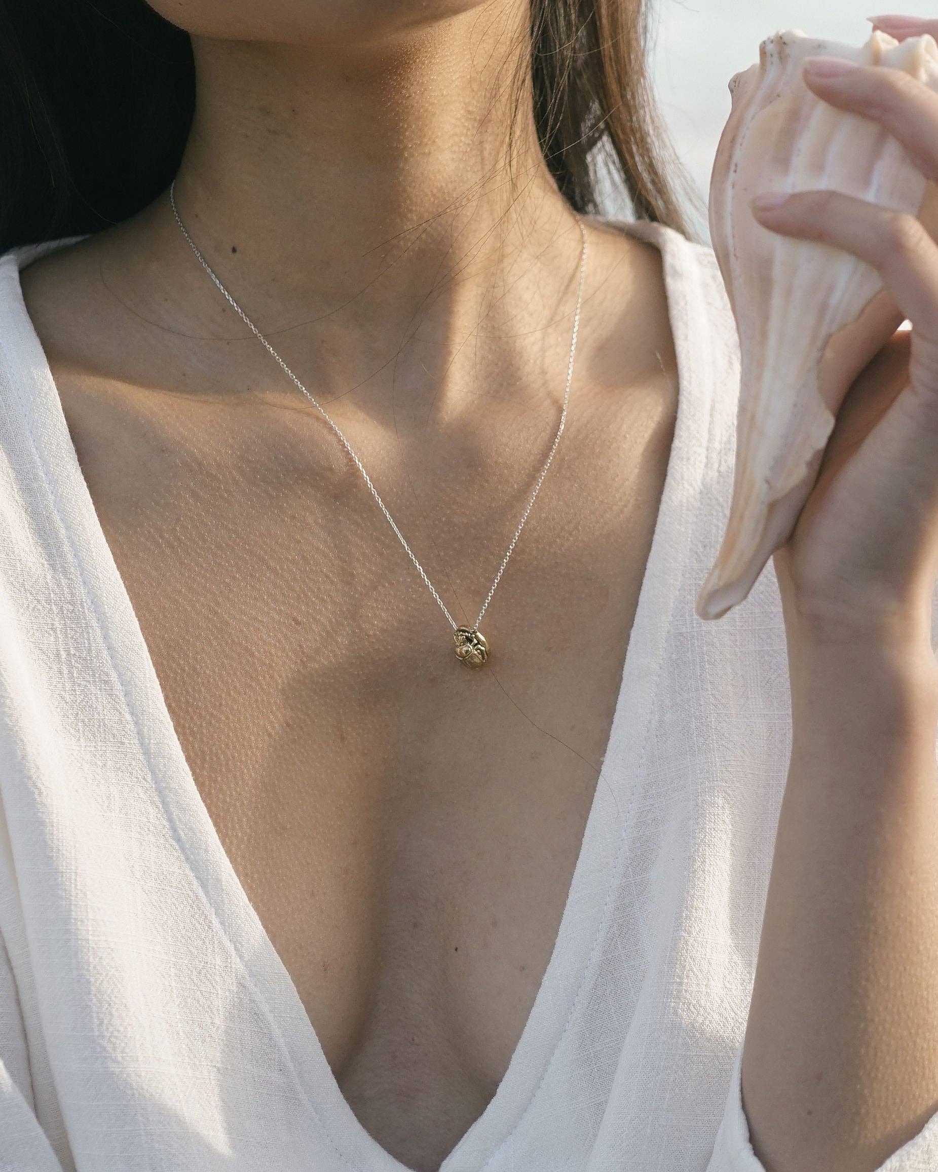 A close-up lifestyle shot of a model wearing a delicate gold necklace with a small, textured geometric pendant. The model is wearing a white V-neck top and holding a large conch shell against a bright, sunlit background.