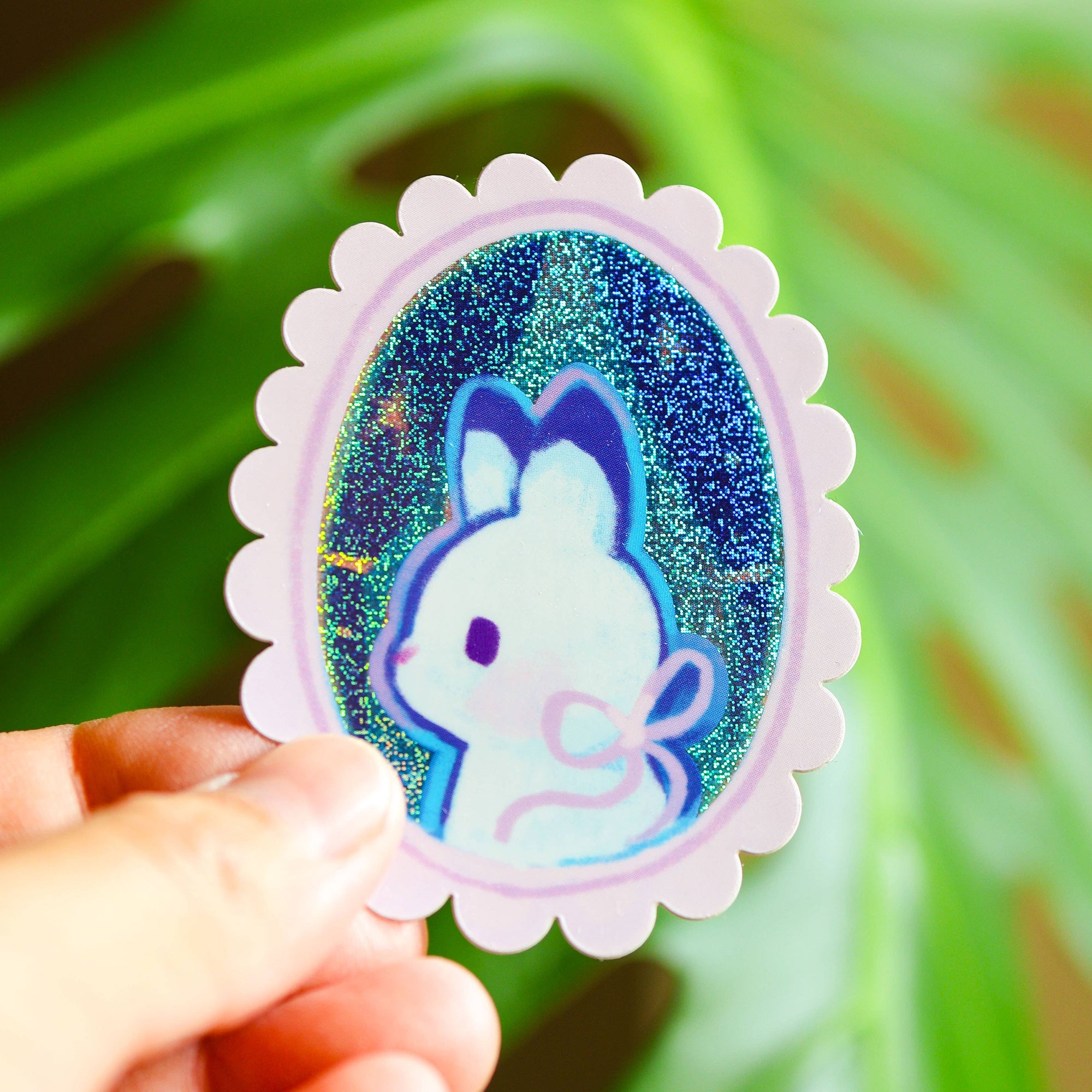 Holographic sticker with a rabbit design held by a hand against a green leafy background