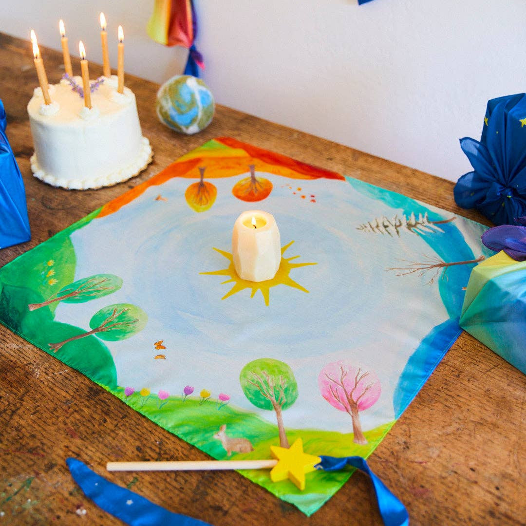 Decorative sheet with a landscape design on a wooden surface, surrounded by candles and a cake.