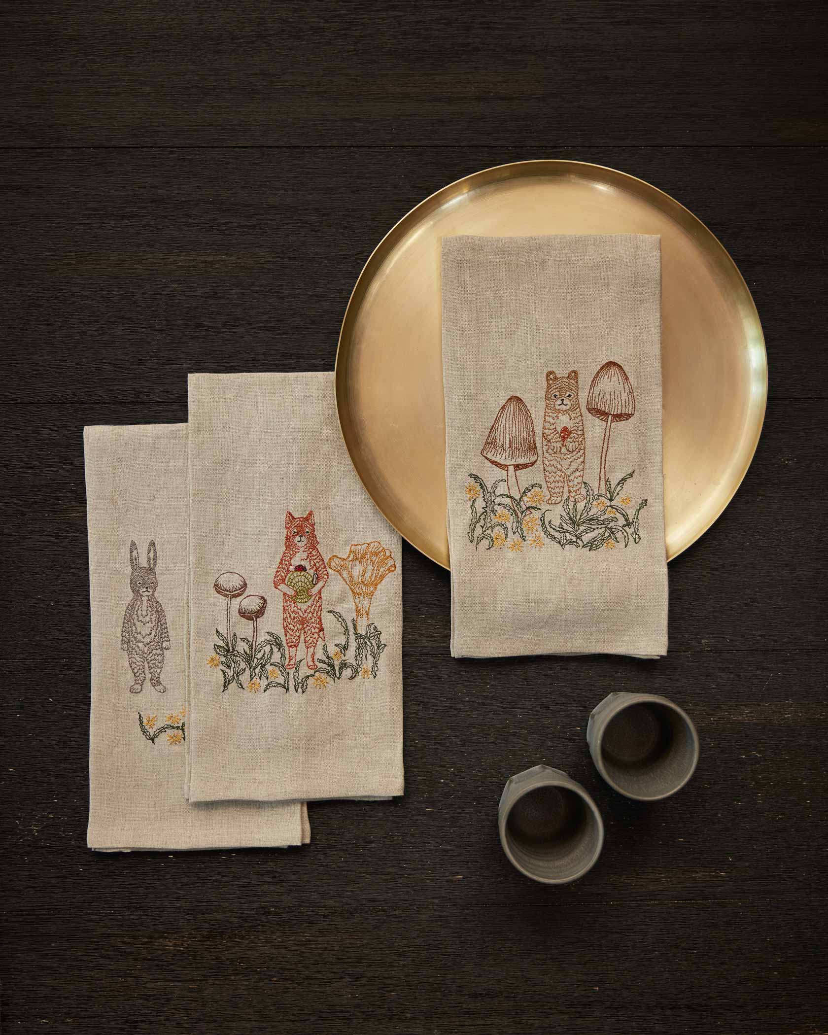 Set of embroidered towels with animal and mushroom designs on a dark surface with a gold plate and ceramic items.