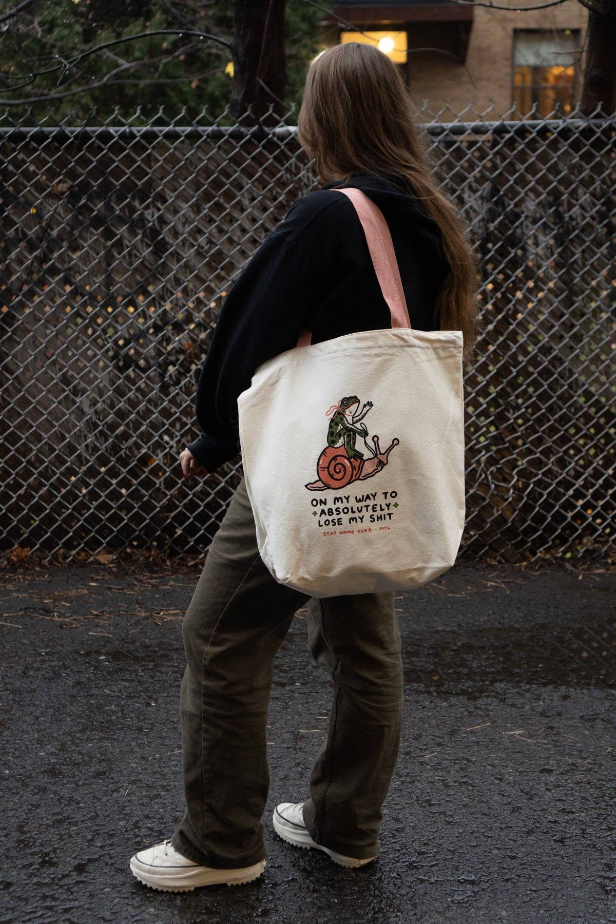 Person holding a tote bag with a graphic design and text, standing in front of a chain-link fence.