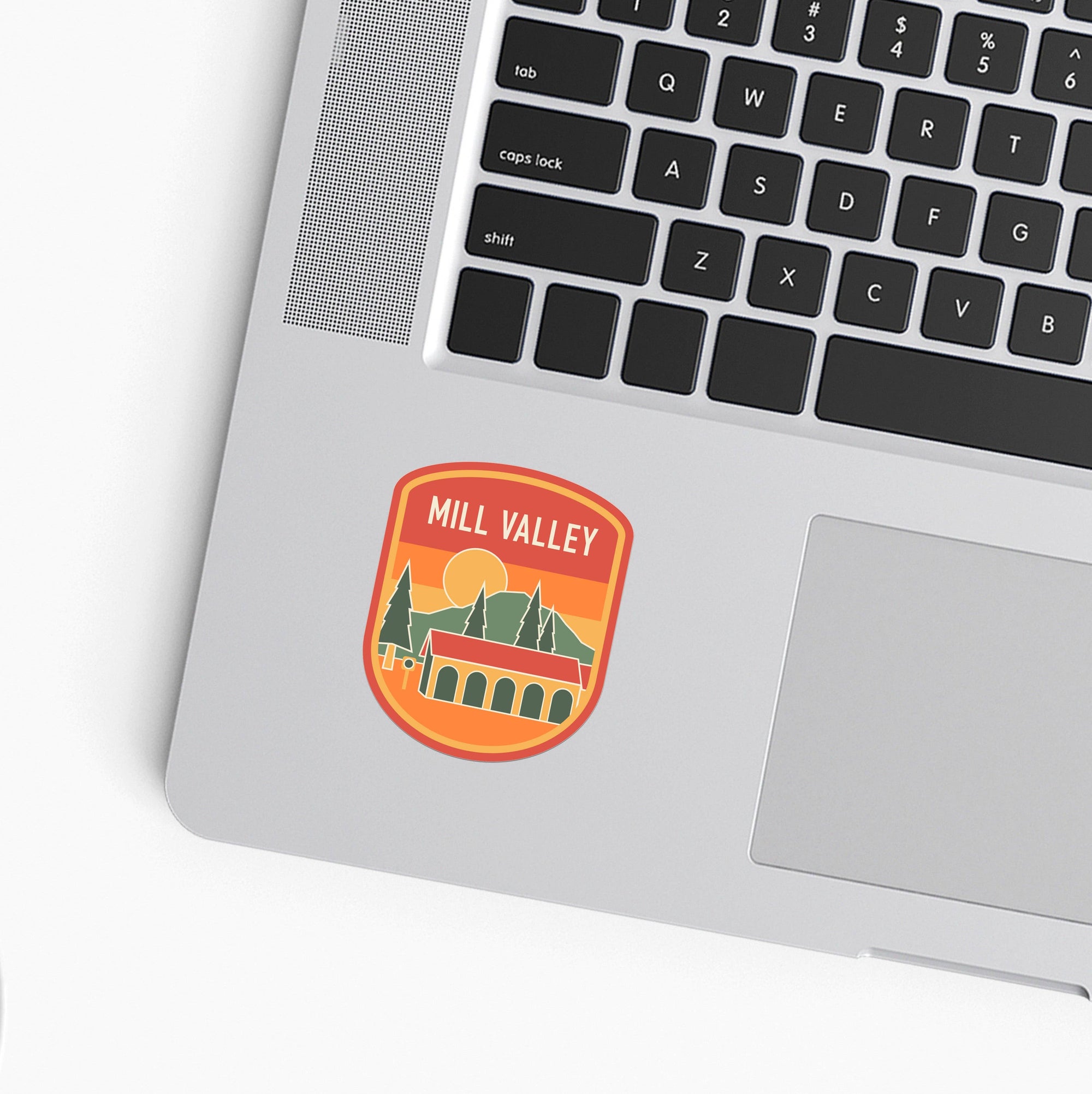A round sticker with a retro Mill Valley landscape design, shown applied to the palm rest of a silver laptop on a white surface.
