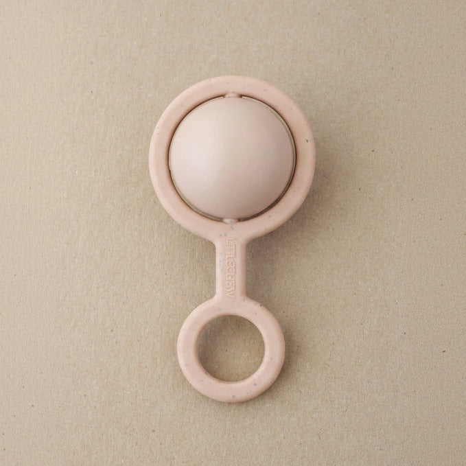 A pale pink baby teething ring toy with a smooth oval ball inside, photographed on a textured beige background.