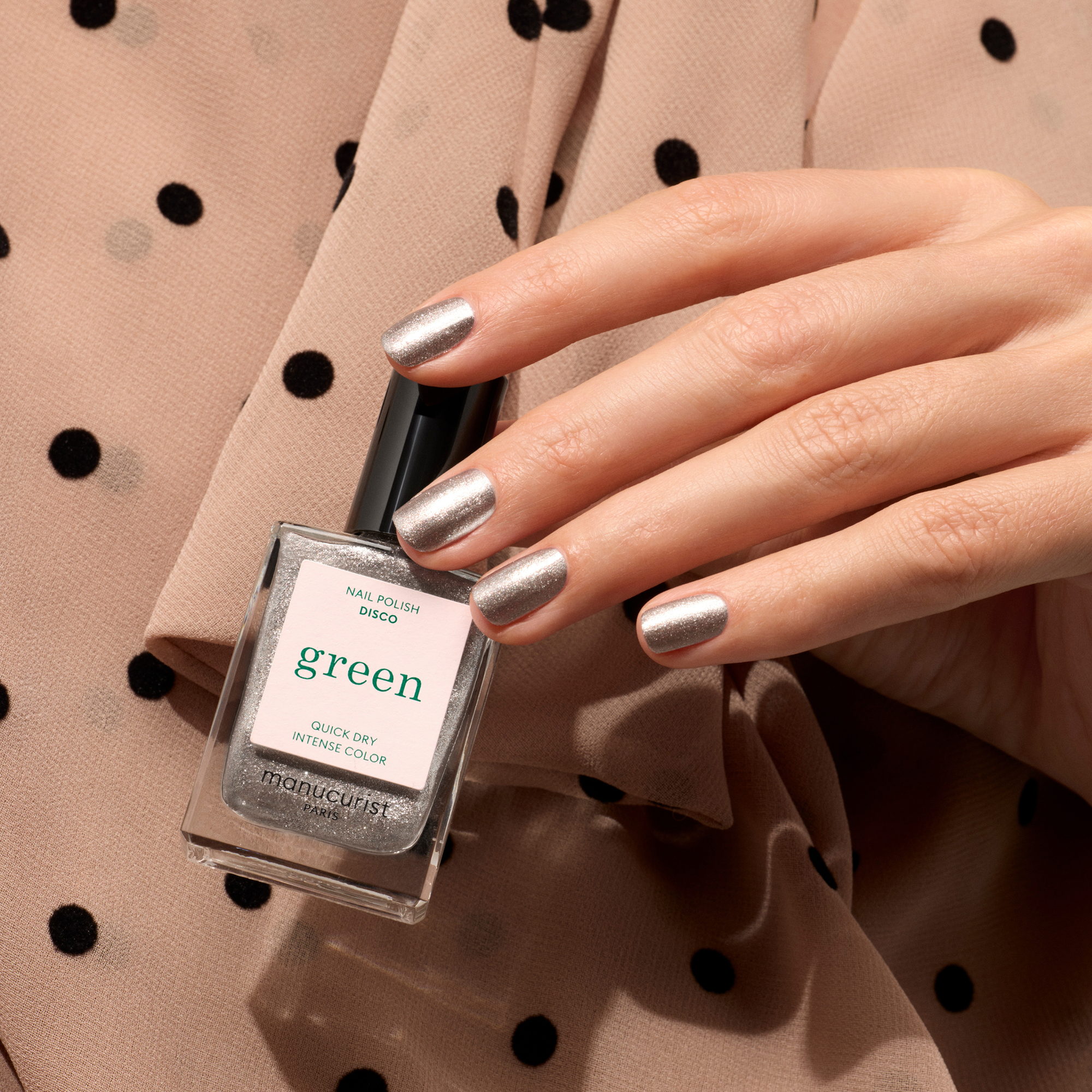 A hand with metallic silver nails holds a bottle of Indigo Girls 'green' nail polish against a beige polka-dot fabric background.