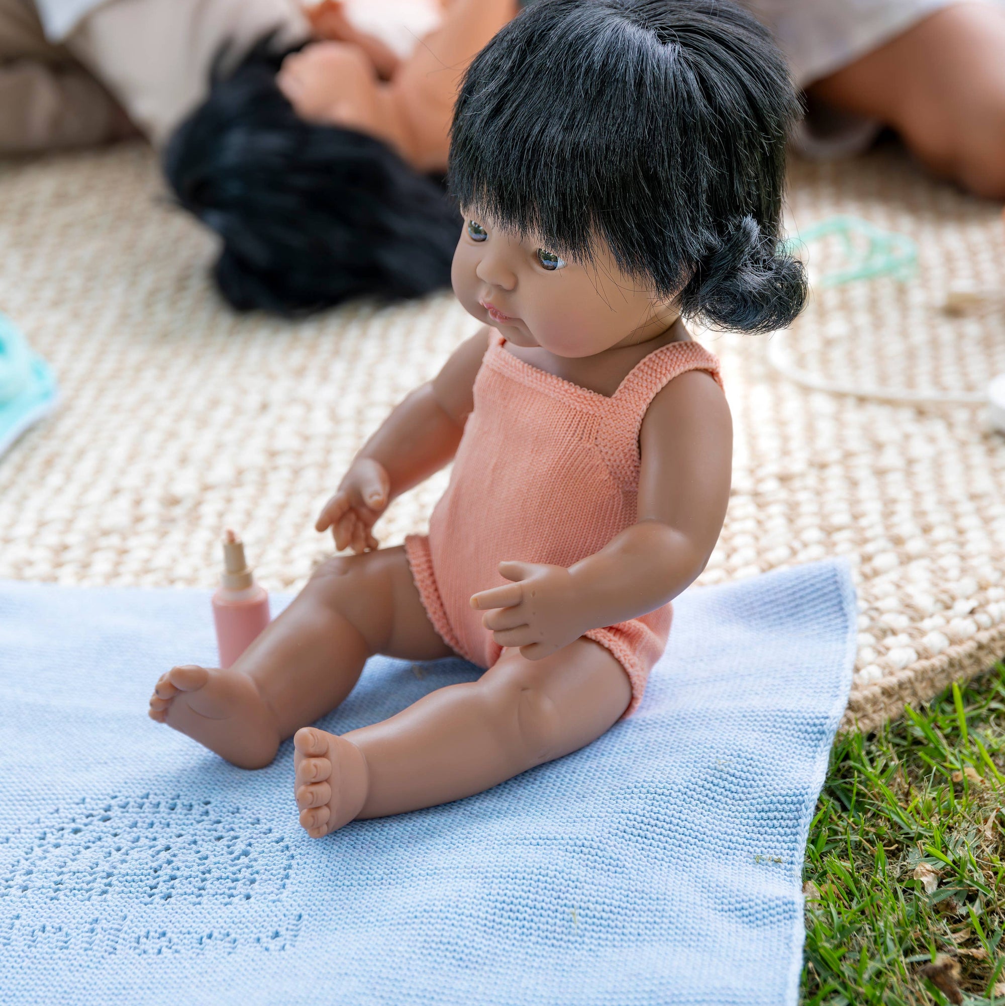 Child's doll with black hair and pink outfit sitting on a blue blanket outdoors.