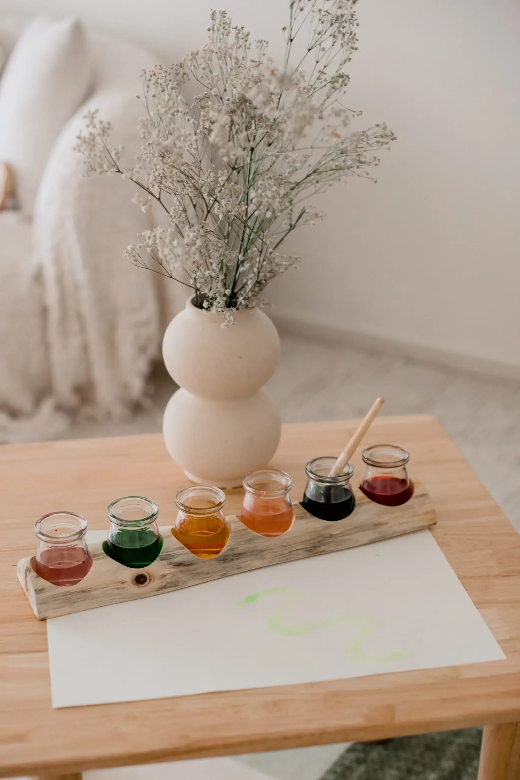Set of small glass containers with colorful liquids on a wooden board, placed on a table with a vase and flowers in the background.