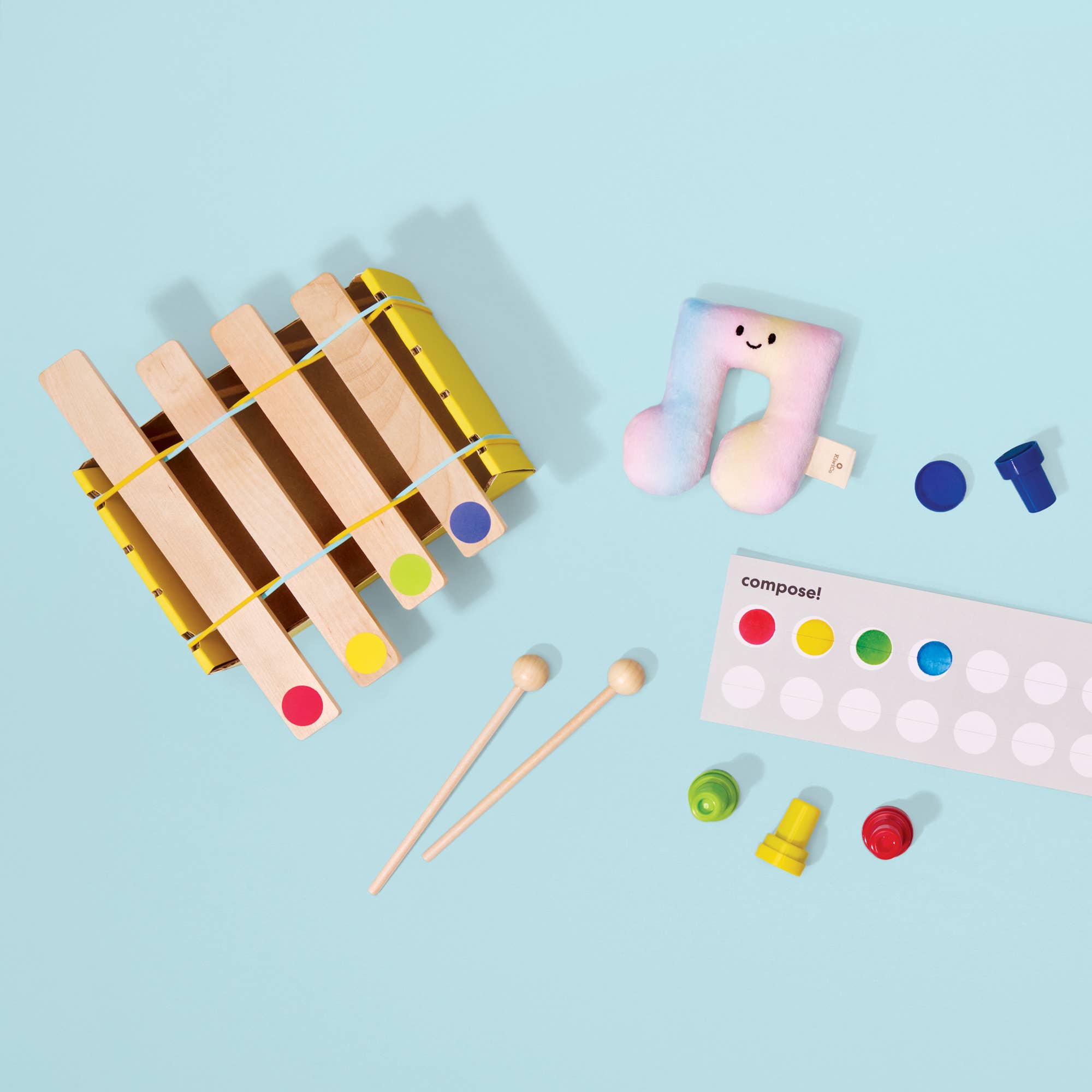 A children's music toy set arranged on a light blue background, including a wooden xylophone, two wooden mallets, a pastel music note pillow, colored percussion pieces, and a white labeled board.