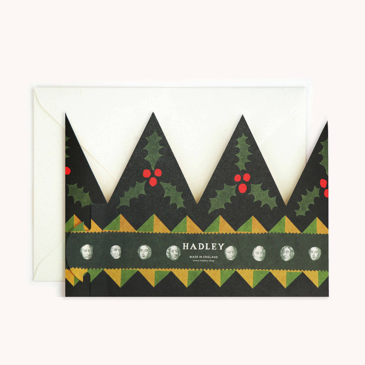 A Christmas-themed greeting card with a crown design, featuring green holly leaves and red berries on a black background, with a white envelope behind it on a light beige background.