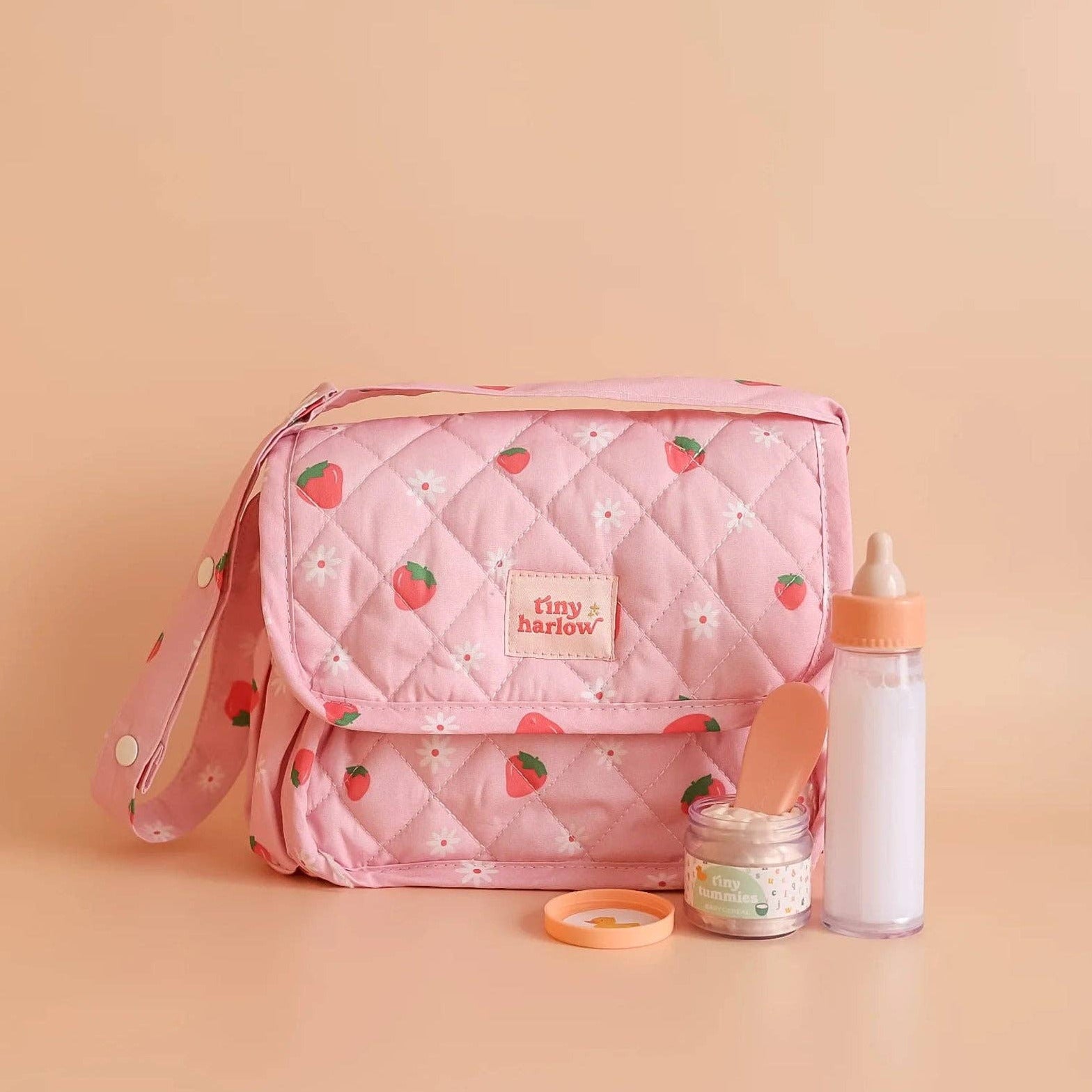 Pink quilted diaper bag with baby doll feeding items on a beige background