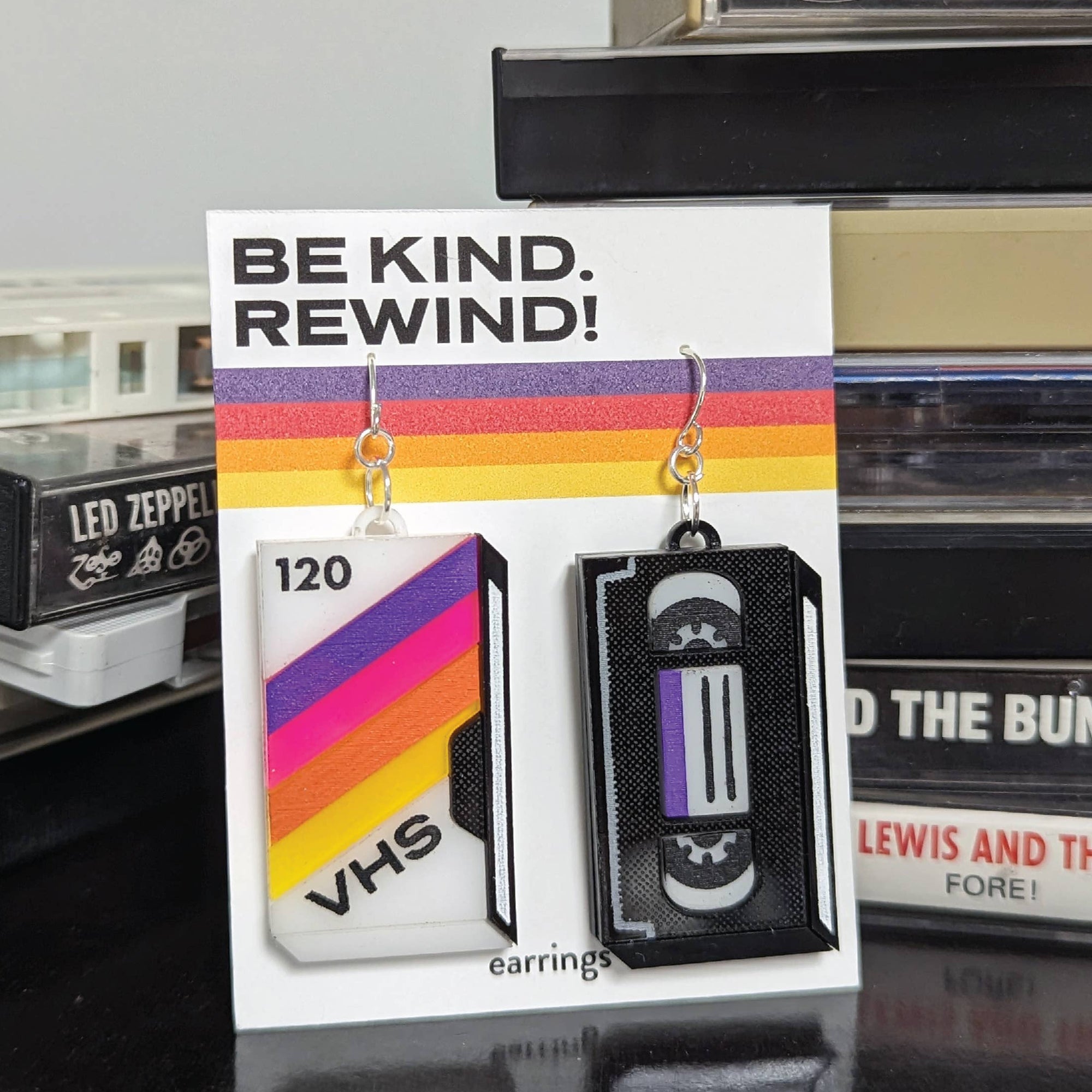 VHS tape earrings on a card with 'Be Kind. Rewind!' text, displayed against a cassette tape background.