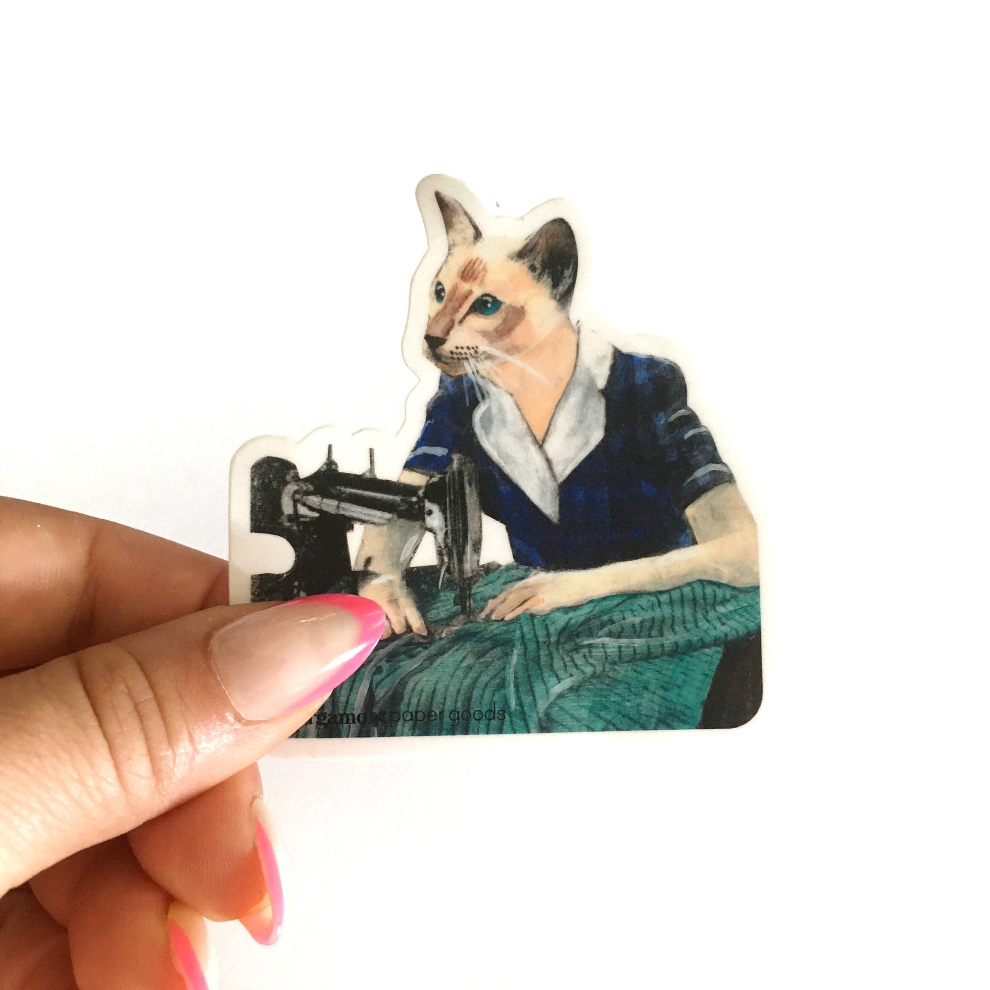 Sticker of a cat operating a sewing machine held by a hand with pink nail polish.