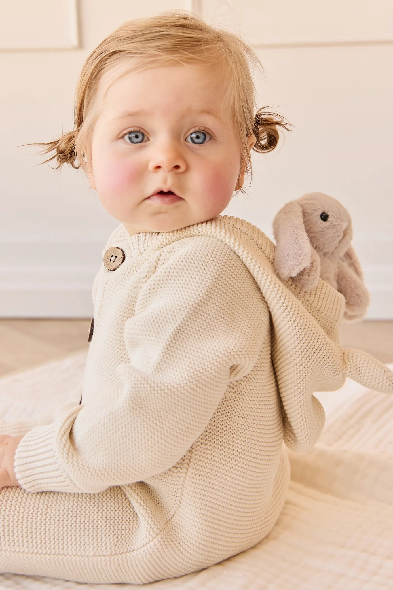 Baby in a beige knitted outfit with bunny ears sitting on a light-colored blanket.