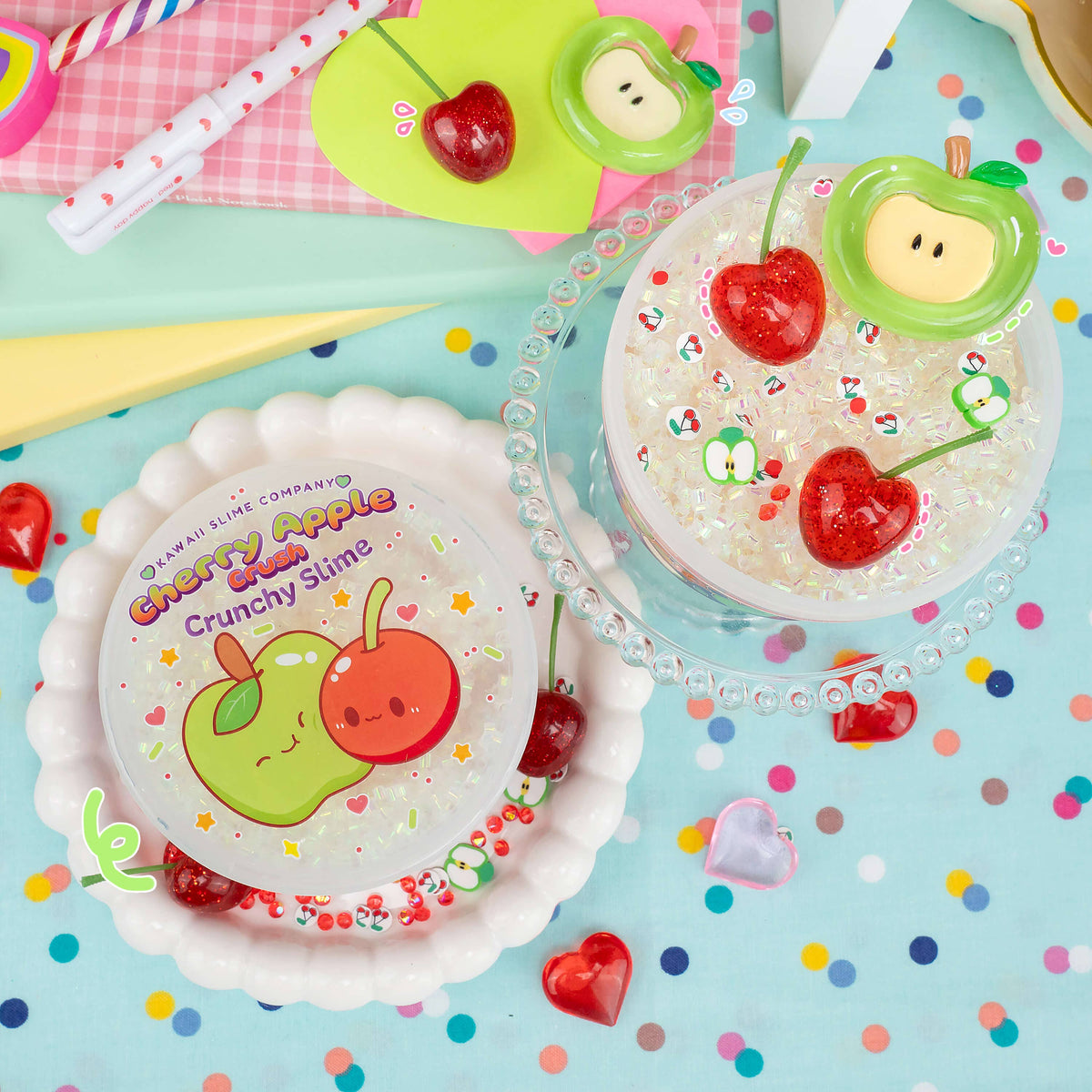 Two cups of fruit-themed crunchy slime displayed on a colorful, polka-dot background. One cup features a lid decorated with a green and red cartoon apple illustration, and the other is open, showing clear slime filled with red heart-shaped candies and small green apple pieces.