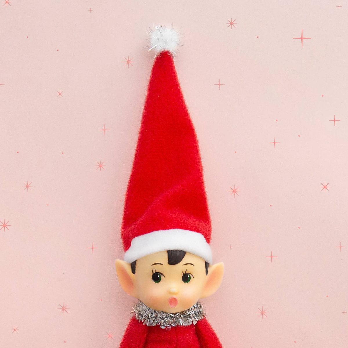 Elf on the Shelf with a red hat and scarf on a pink background