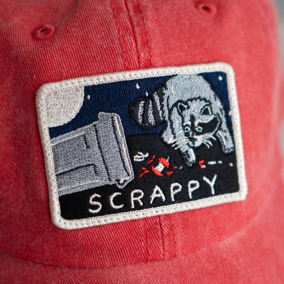 Red cap with a patch featuring a racoon and the word 'SCRAPPY'.