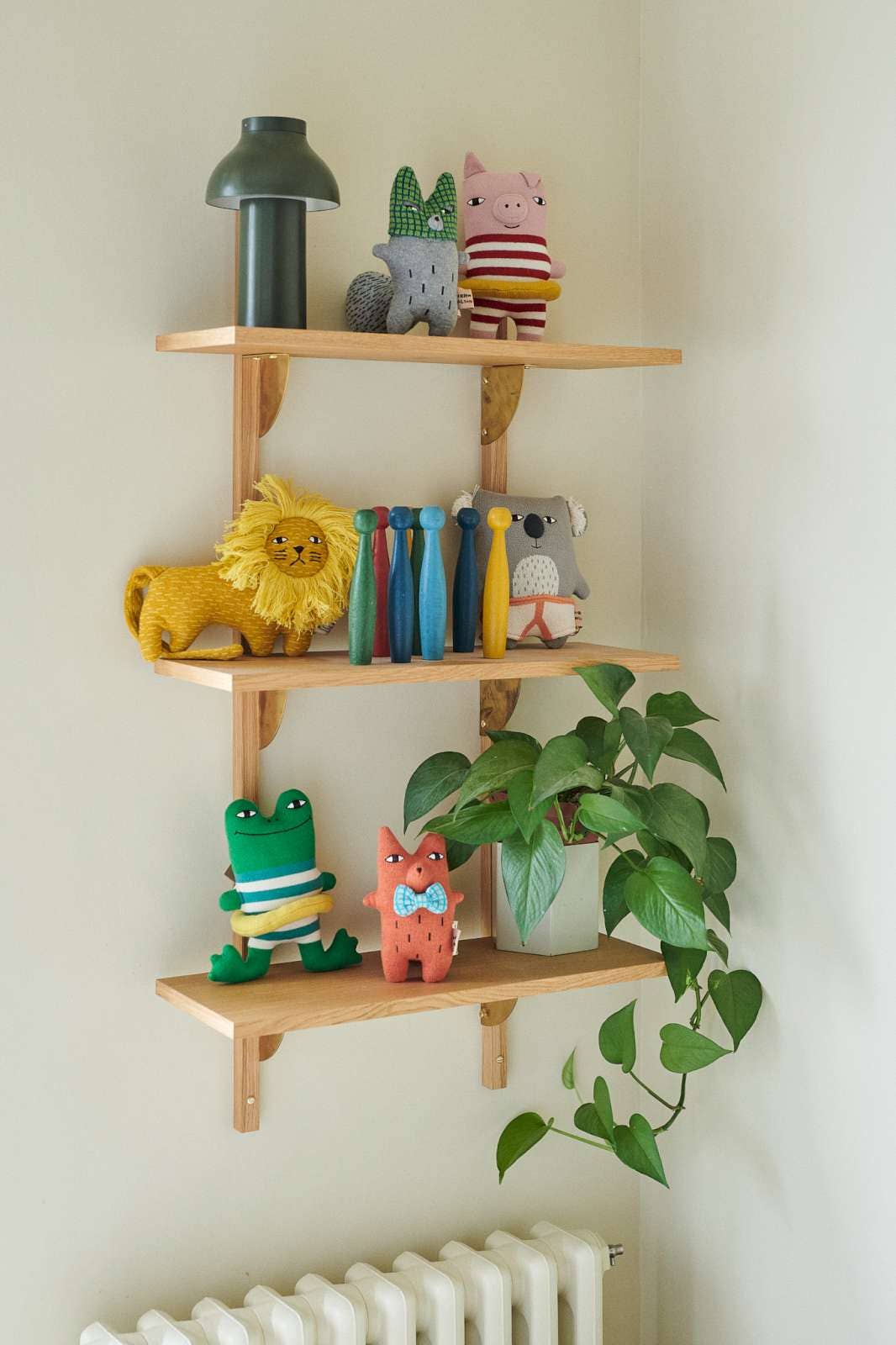 Three light natural wood floating shelves mounted on a white wall, styled with colorful plush toys, a green lamp, a potted green plant, and a white radiator below.
