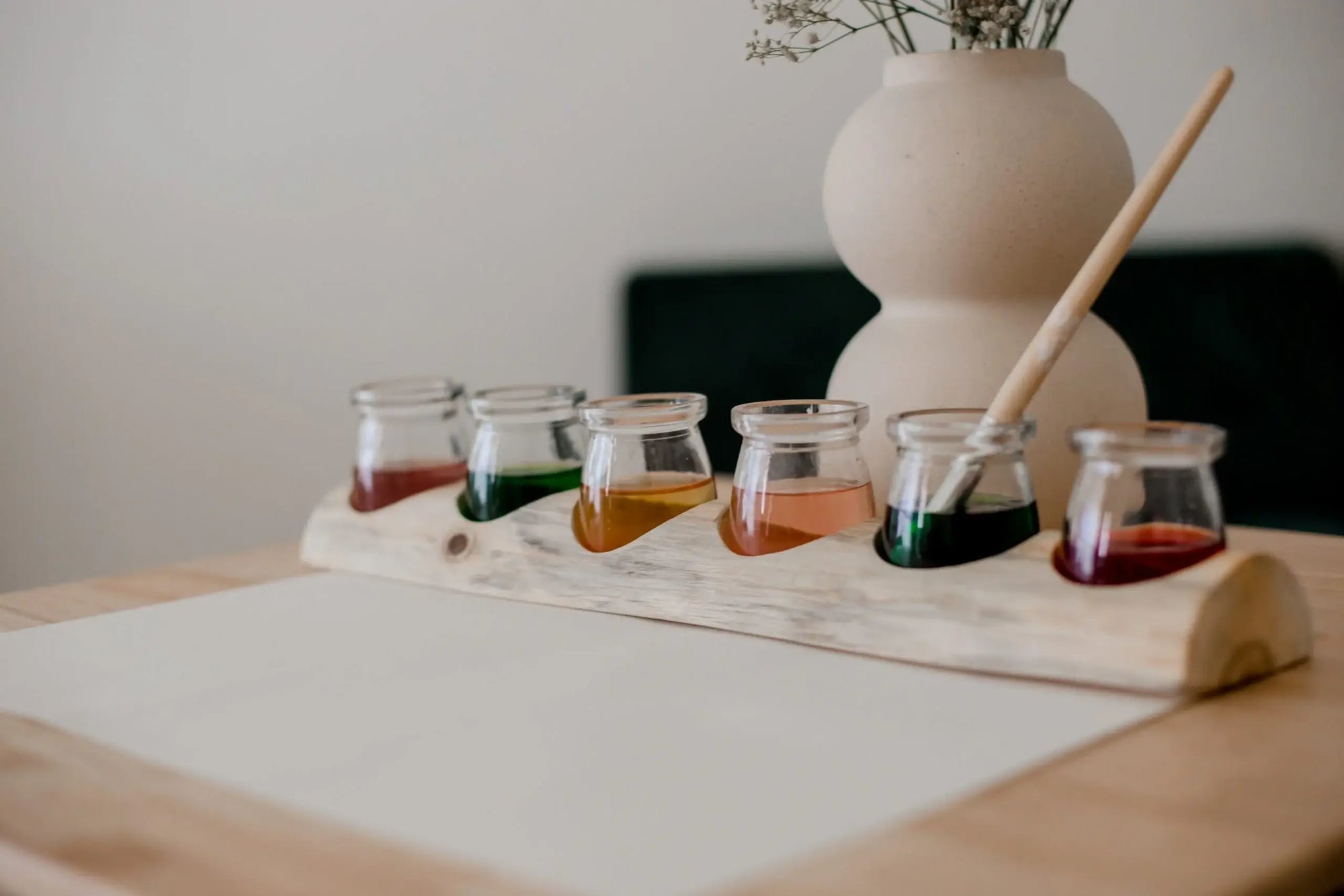 Set of small glass jars with colorful liquids on a wooden tray, with a vase and brush in the background.