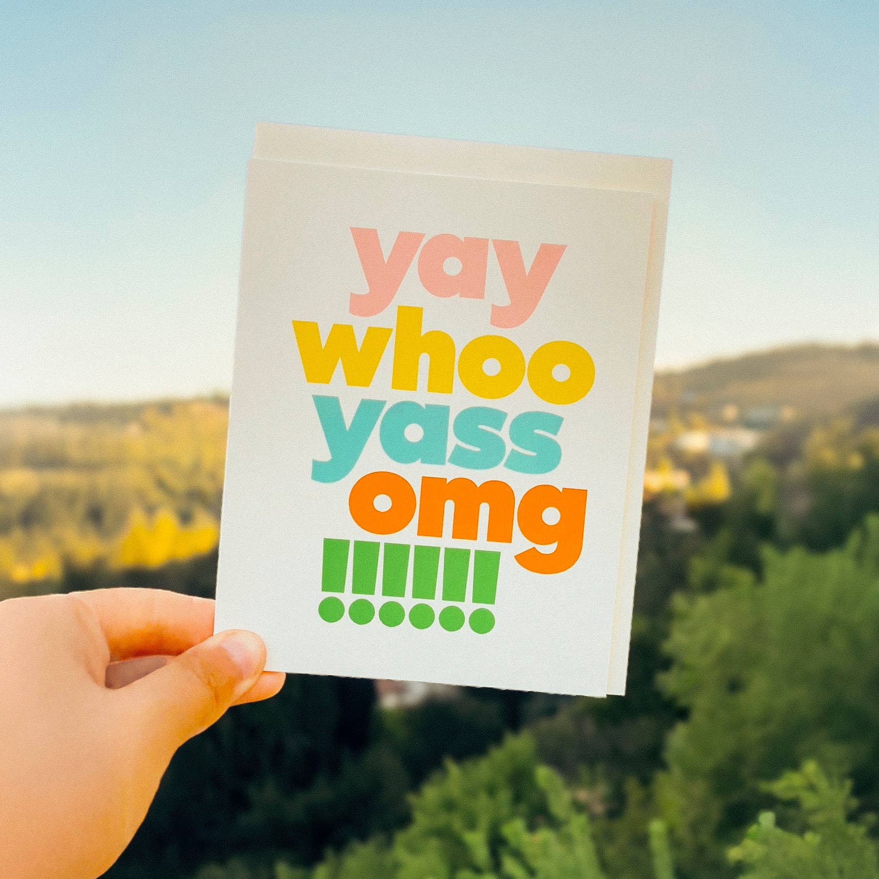 Hand holding a sign with colorful text 'yay whoo yass omg!!!!!!' against a scenic background.