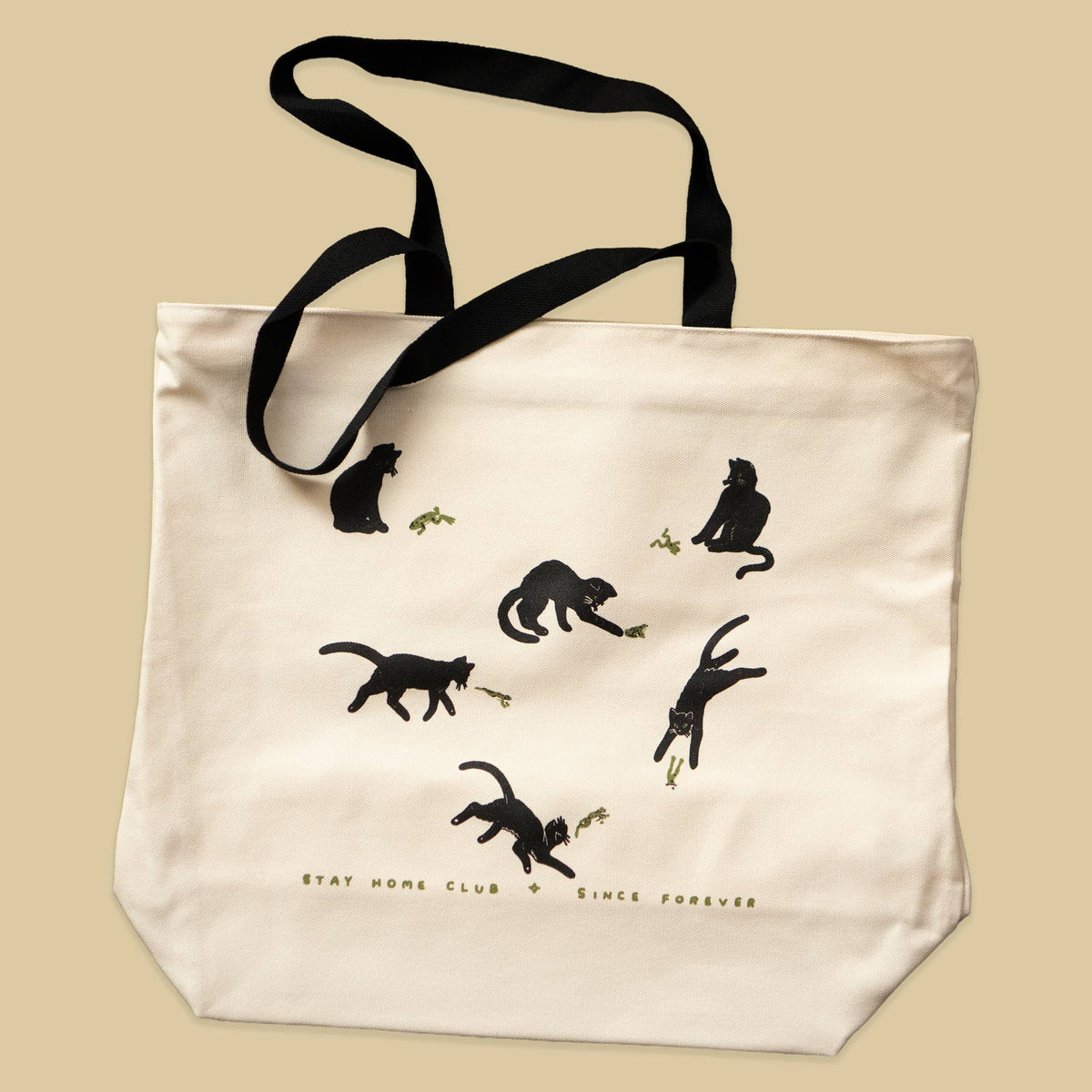 An off-white canvas tote bag with black handles, featuring a playful black cat silhouette illustration and the text 'STAY HOME CLUB + SINCE FOREVER' on a solid beige background.