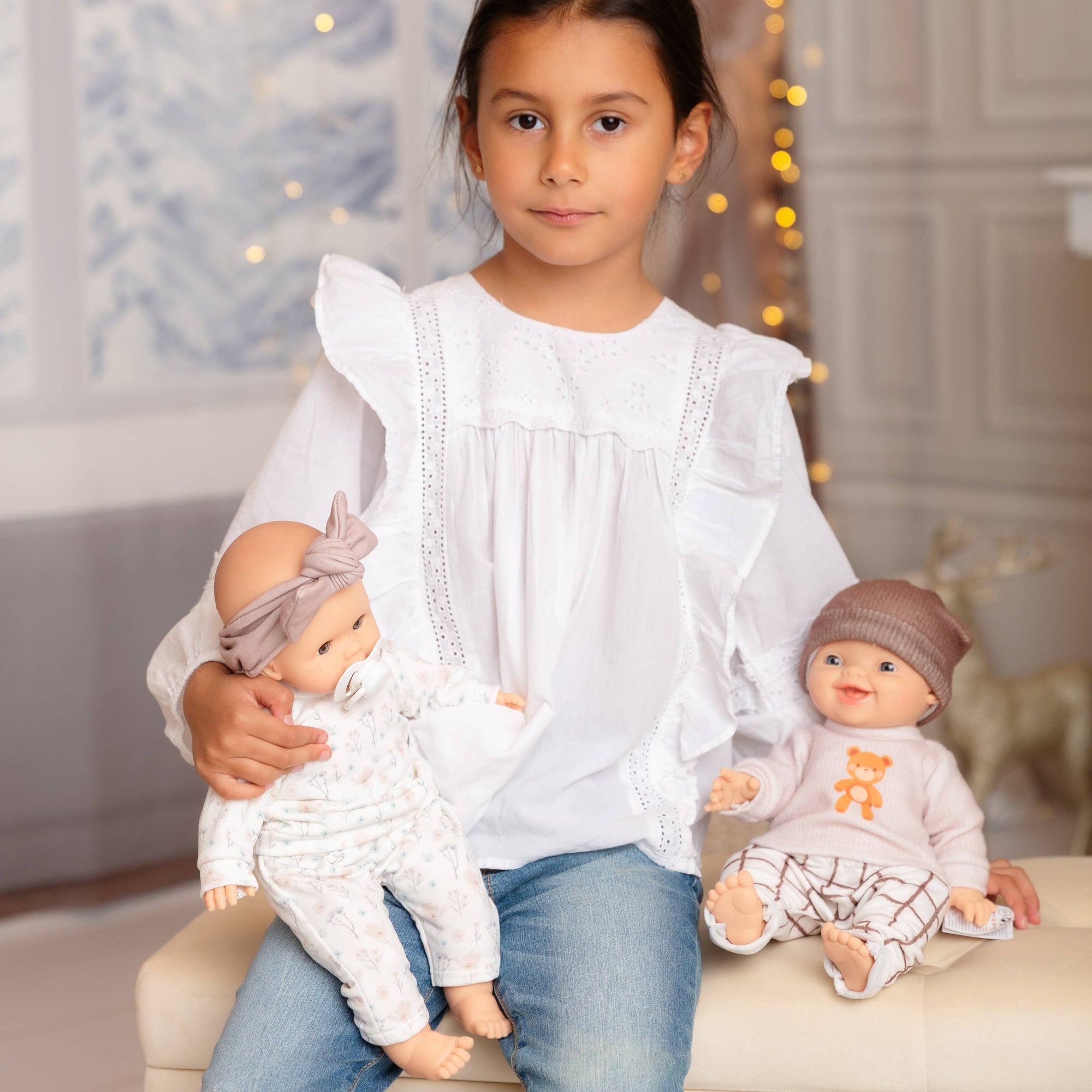 Young girl holding two dolls in a decorated room with lights and curtains.