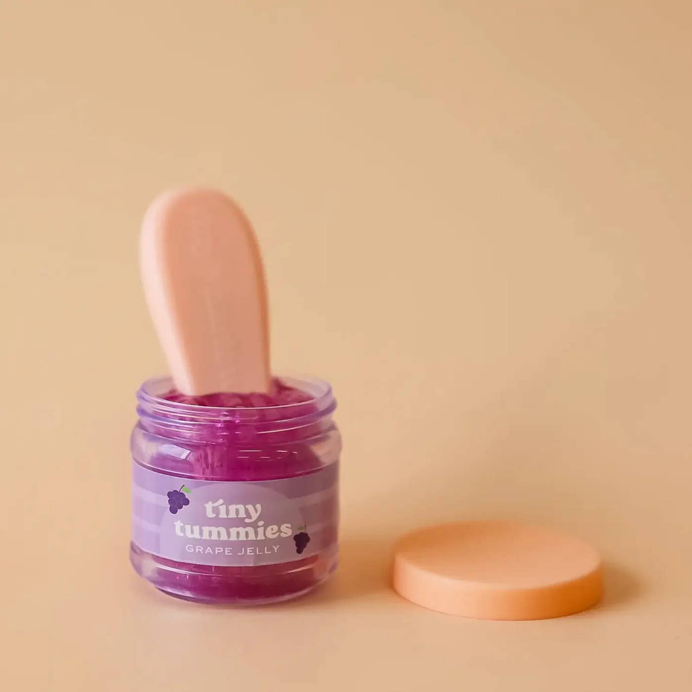 Jar of purple jelly with a pink spoon on a beige background, featuring 'tiny tummies' branding.