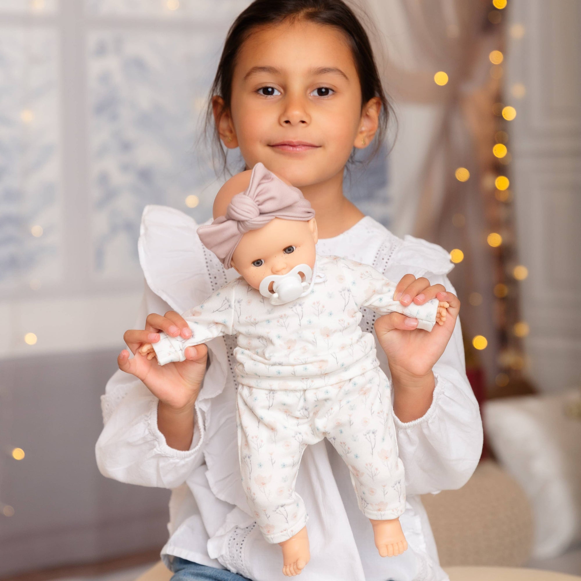 Young girl holding a baby doll with a bow in a softly lit room.