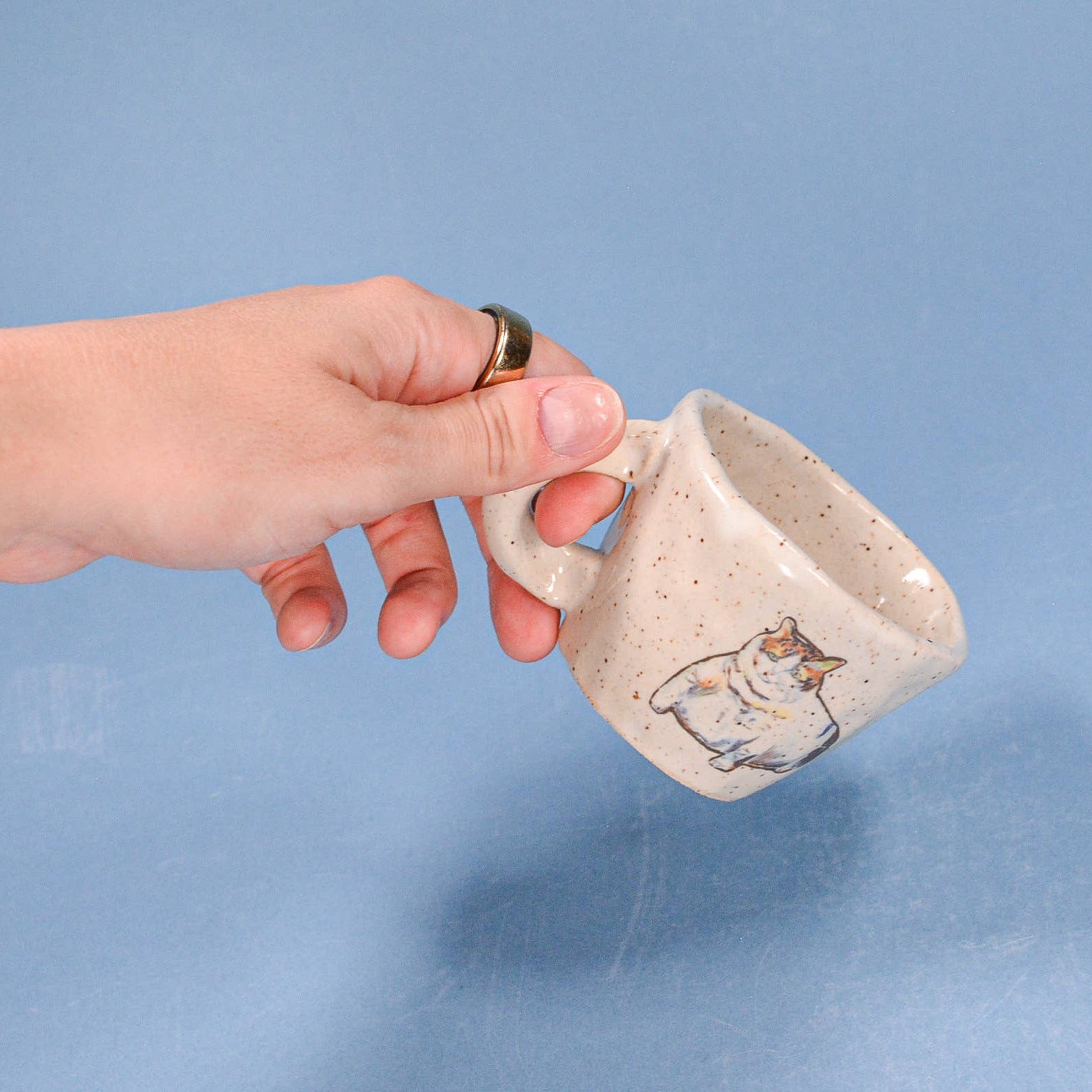 Hand holding a small ceramic mug with a cat design on a blue background