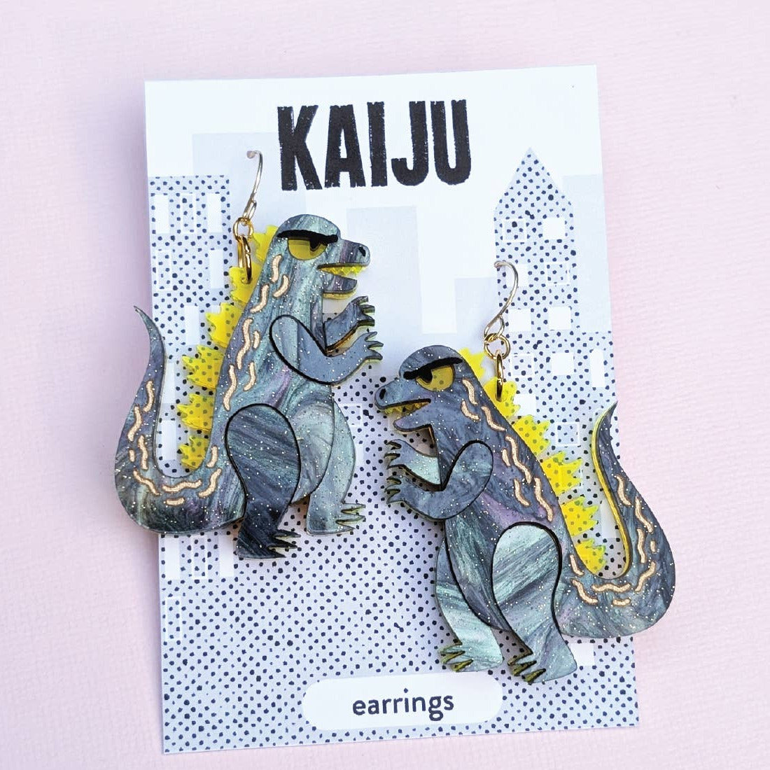 Kaiju-themed earrings on a card with 'Sylvia Draws!' branding against a light pink background.