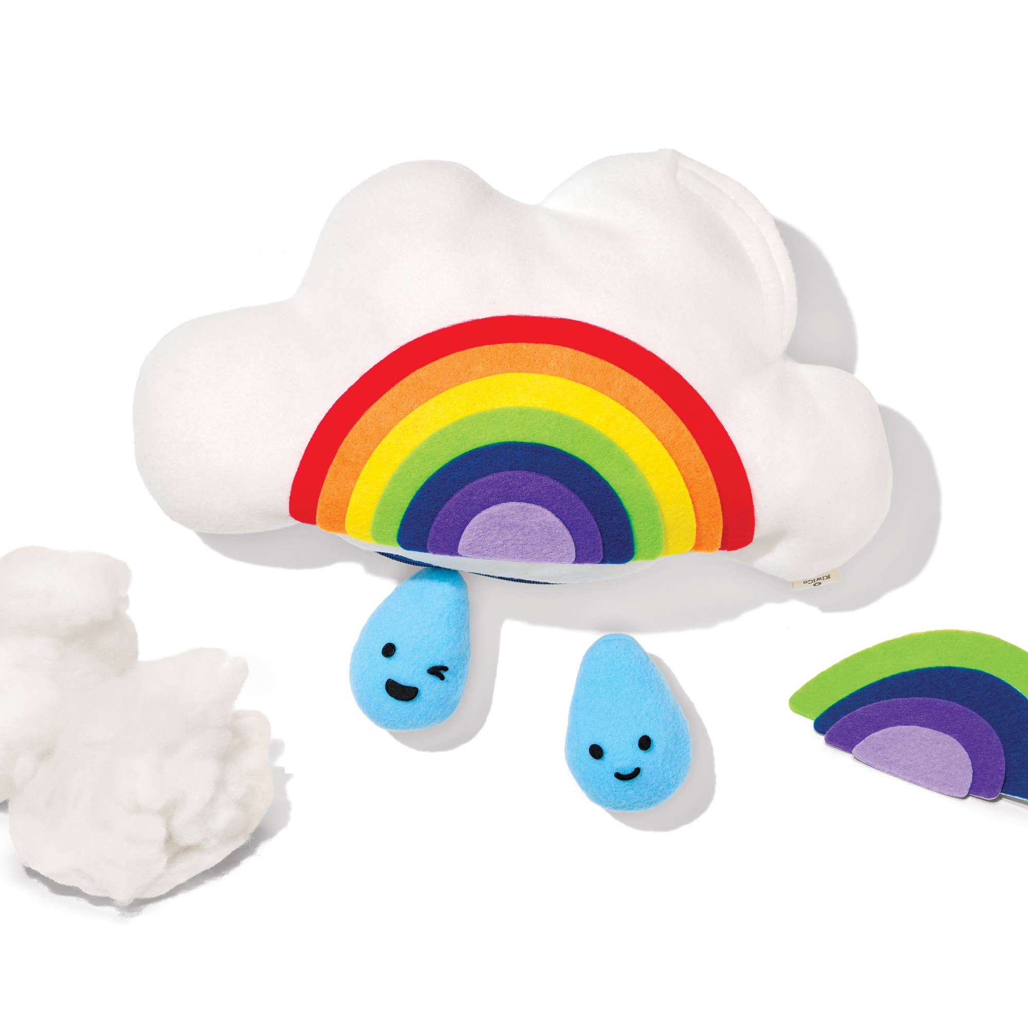A white plush cloud toy with a colorful felt rainbow attached across the front and two small blue teardrop shapes with smiling faces hanging below. A partial matching rainbow and a clump of white stuffing are visible on a plain white background.