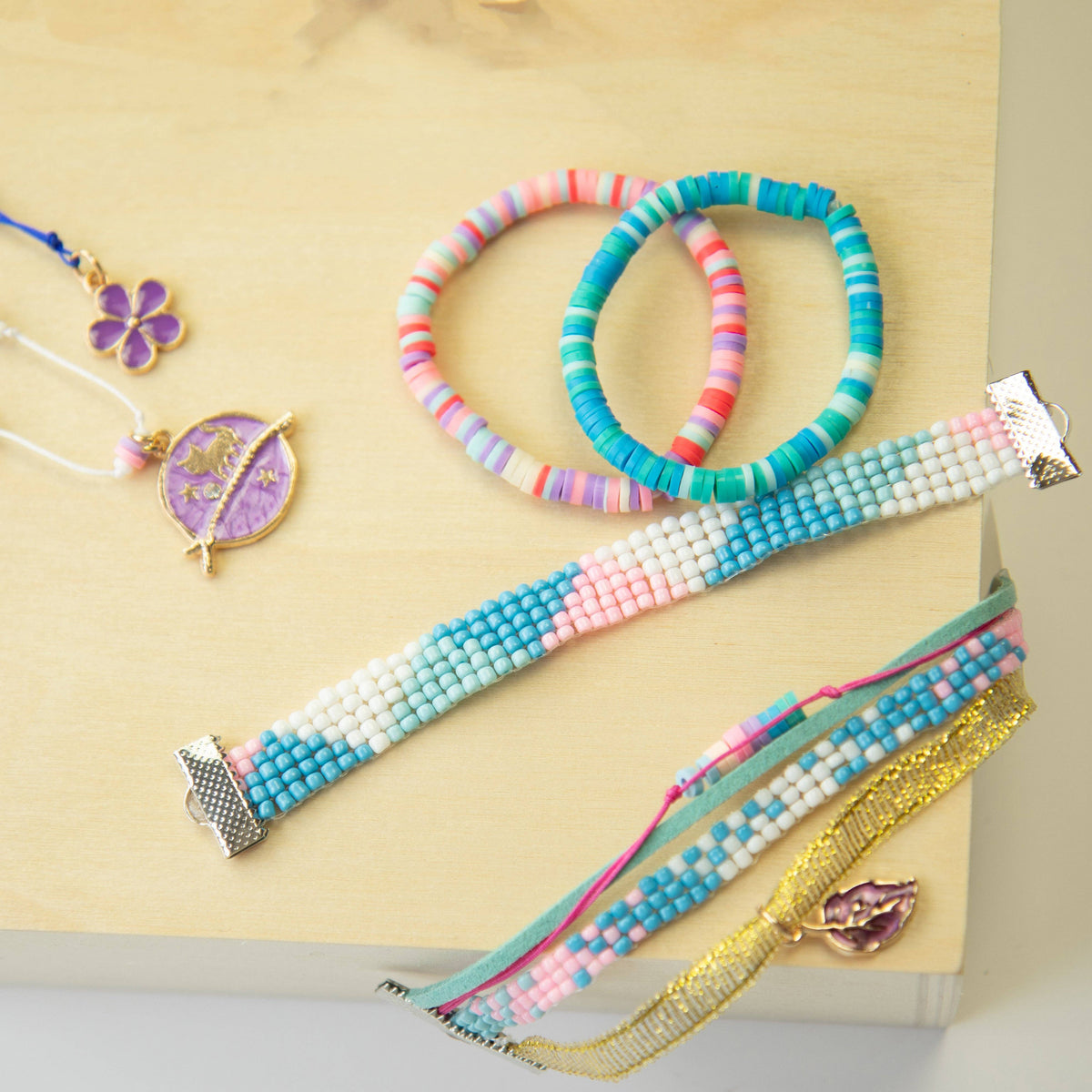 A styled flat-lay of handmade beaded jewelry on a light wood background, featuring two beaded bracelets, two beaded rings, and two beaded necklaces with purple and gold charms.