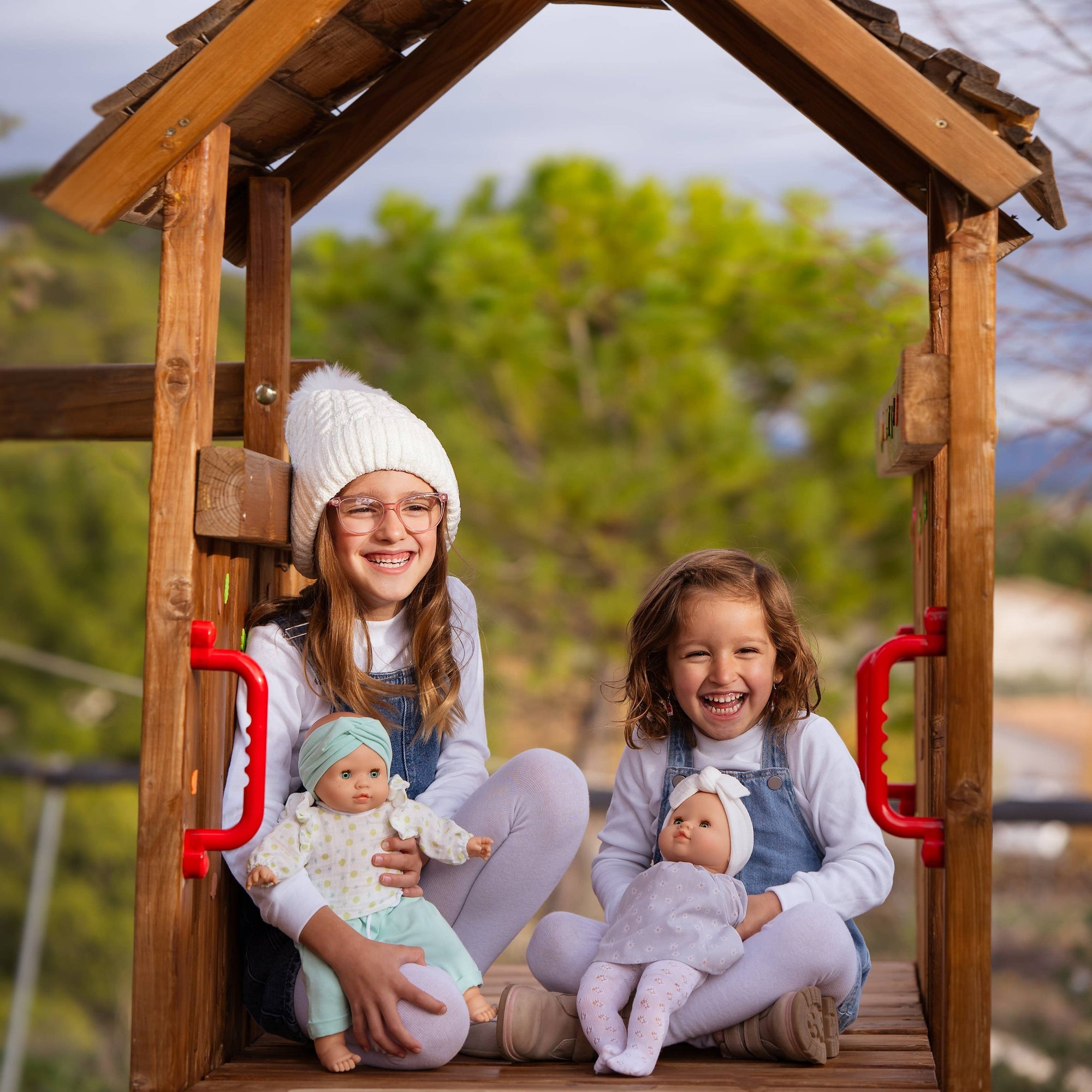 Two children sitting in a wooden playhouse with dolls, surrounded by greenery.