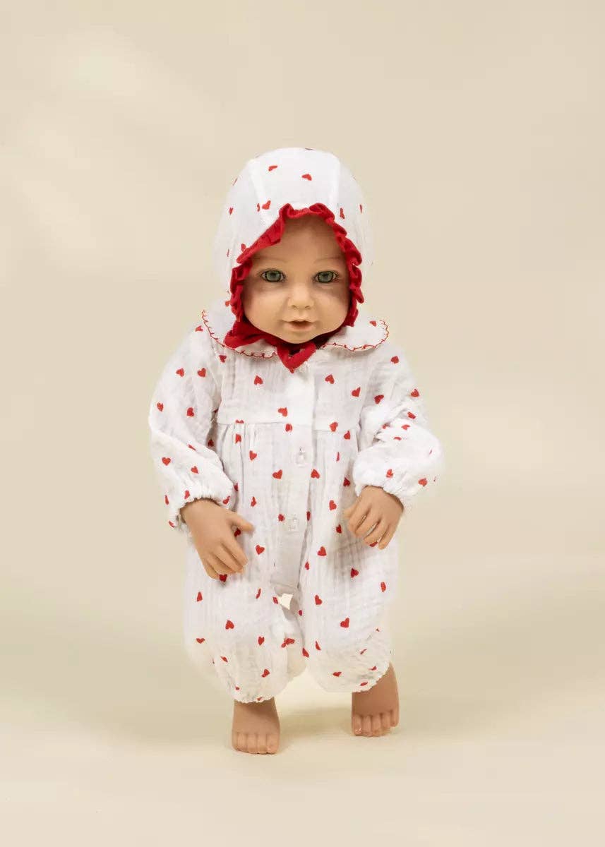 A baby doll wearing a white muslin one-piece jumpsuit with small red hearts and a matching hood with red ruffle trim, standing against a plain beige background.