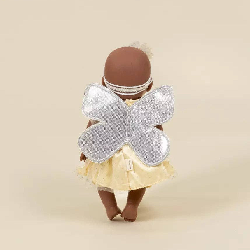 A doll viewed from behind wearing a silver glitter butterfly wing accessory, a pale yellow skirt, and a white beaded headband against a solid beige background.