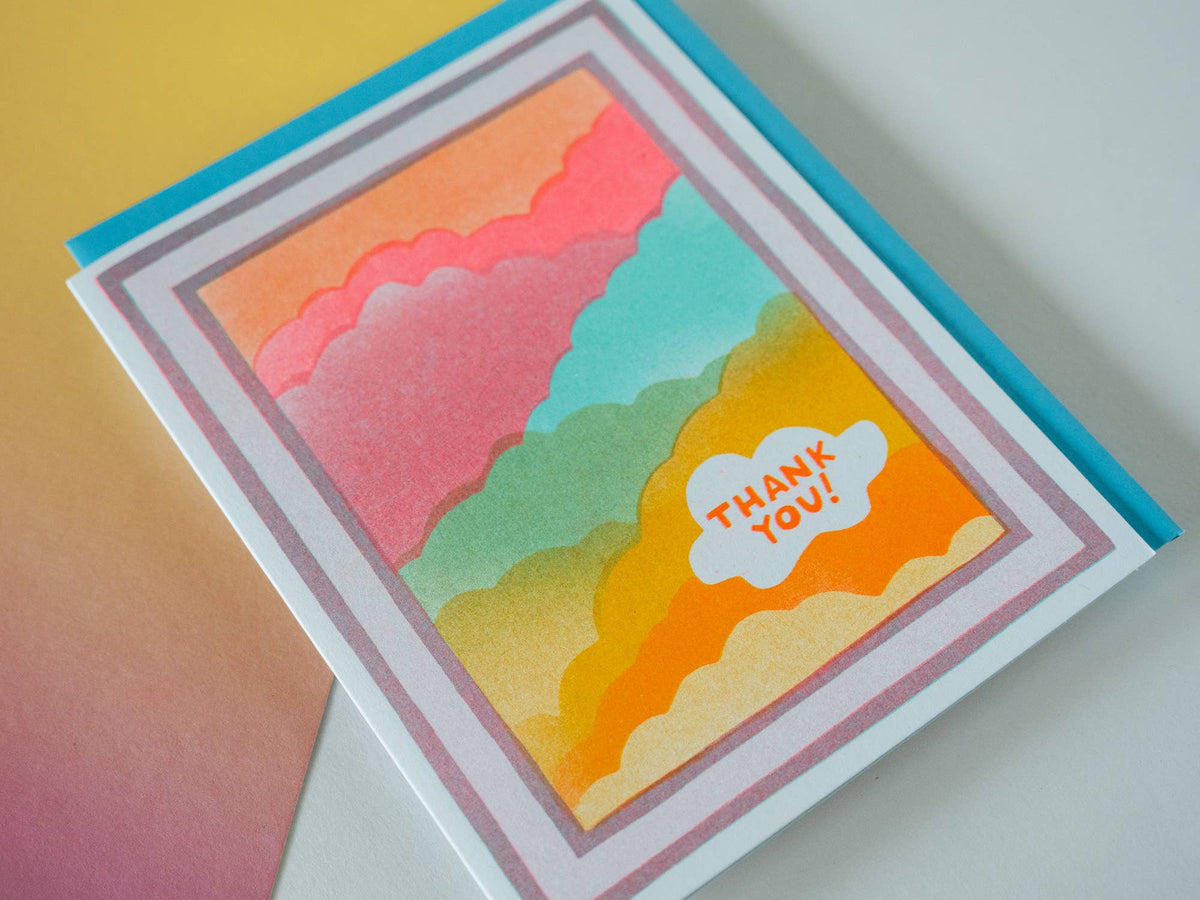 A handmade greeting card featuring a colorful layered cloud pattern in orange, pink, teal, green, and yellow, with a white cloud stamp that reads 'THANK YOU!' in orange. The card is layered over a bright blue backing card and photographed on a pastel gradient background.