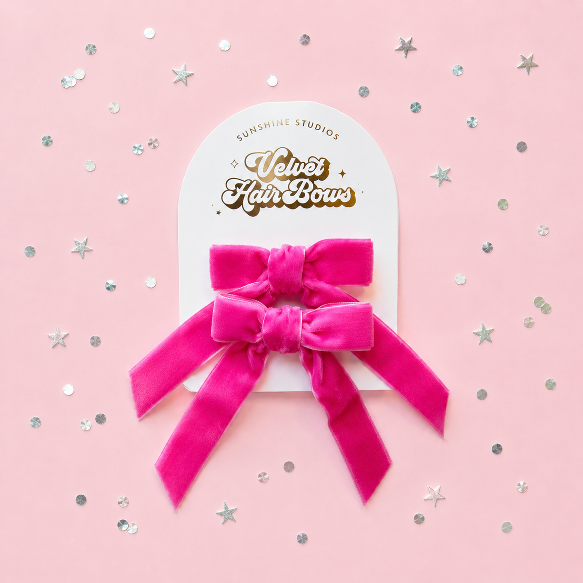 A bright pink velvet hair bow attached to a white display card reading 'Sunshine Studios Velvet Hair Bow,' photographed on a light pink background with scattered silver star and dot confetti.