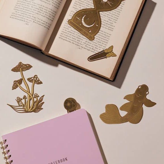 Open book with decorative bookmarks shaped like mushrooms, a sword, and a fish, next to a pink notebook on a light surface.
