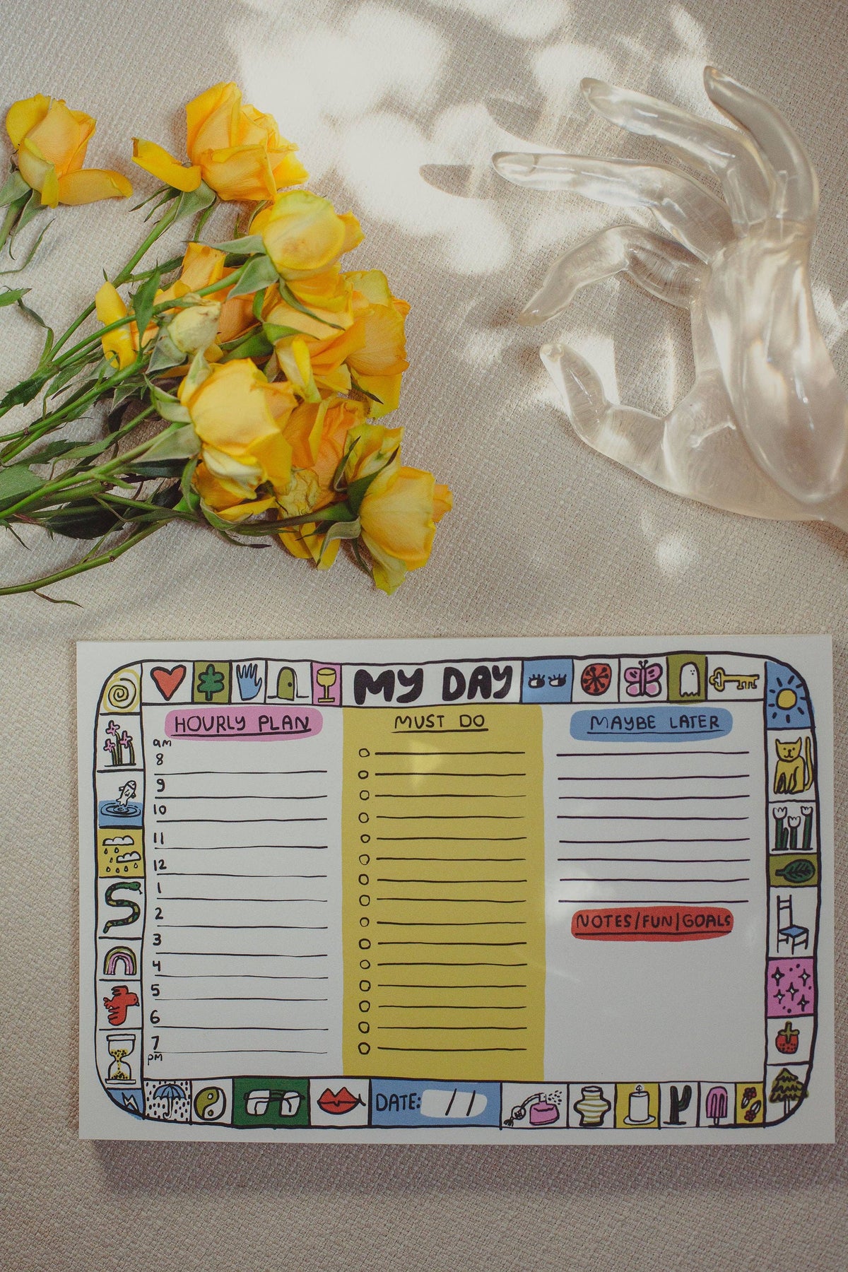 A weekly planner notepad titled 'MY DAY' with colorful cartoon border illustrations, laid flat on a beige fabric surface with yellow roses and a clear glass tumbler nearby.